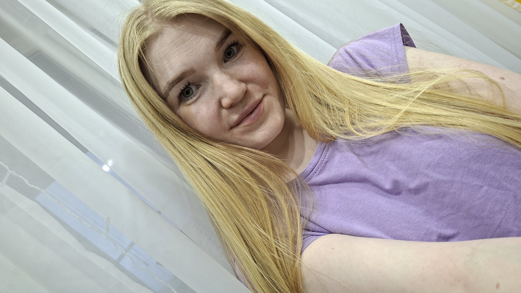 A close-up selfie of a fair-skinned woman with long blonde hair wearing a purple t-shirt is captured indoors. The background reveals a partially visible white curtain or blind, suggesting a window covering. The location, based on contextual clues provided, is in the city of Chaikovsky, Russia.