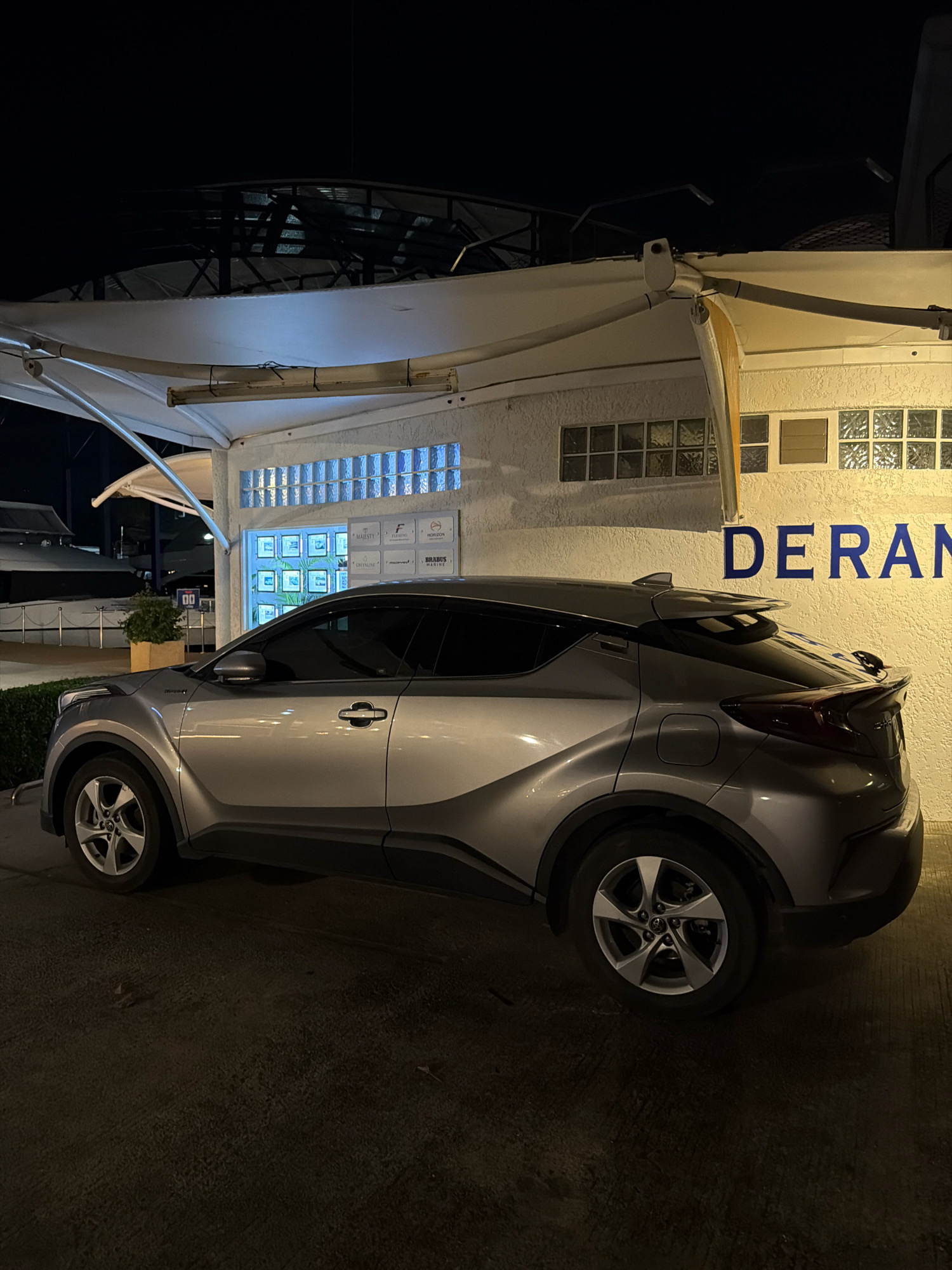 A silver Toyota C-HR is parked outside a building at night in Ko Kaeo, Thailand. The building has a textured white facade with a beige awning extending from the roof. Several small windows with glass blocks are visible above a row of framed pictures. A sign with blue lettering that reads "DERAN" is mounted on the wall. The car is positioned in front of the building, with its side facing the camera. The ground is paved and dark, with some scattered debris. In the background, lights are visible from structures and water, suggesting a marina or waterfront area. The sky is dark, indicating nighttime.