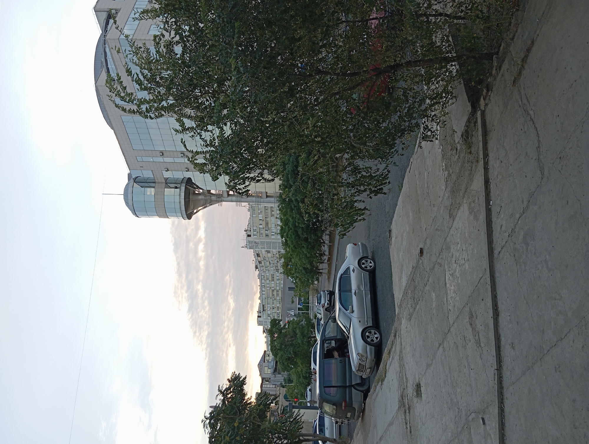The image shows a street scene in Aktau, Kazakhstan, with a focus on a modern glass-fronted office building and parked cars. The building, which has a distinctive curved glass tower section, is situated on the right side of the frame. In the foreground, a silver sedan and a dark-colored minivan are parked along a concrete sidewalk. Beyond these vehicles, a tree with dense green foliage partially obscures the view of the street and other buildings. In the background, several multi-story apartment buildings are visible, indicative of a residential area. The sky is overcast with scattered clouds. The overall impression is an urban environment with a mix of contemporary architecture and older residential blocks, under a pale sky.