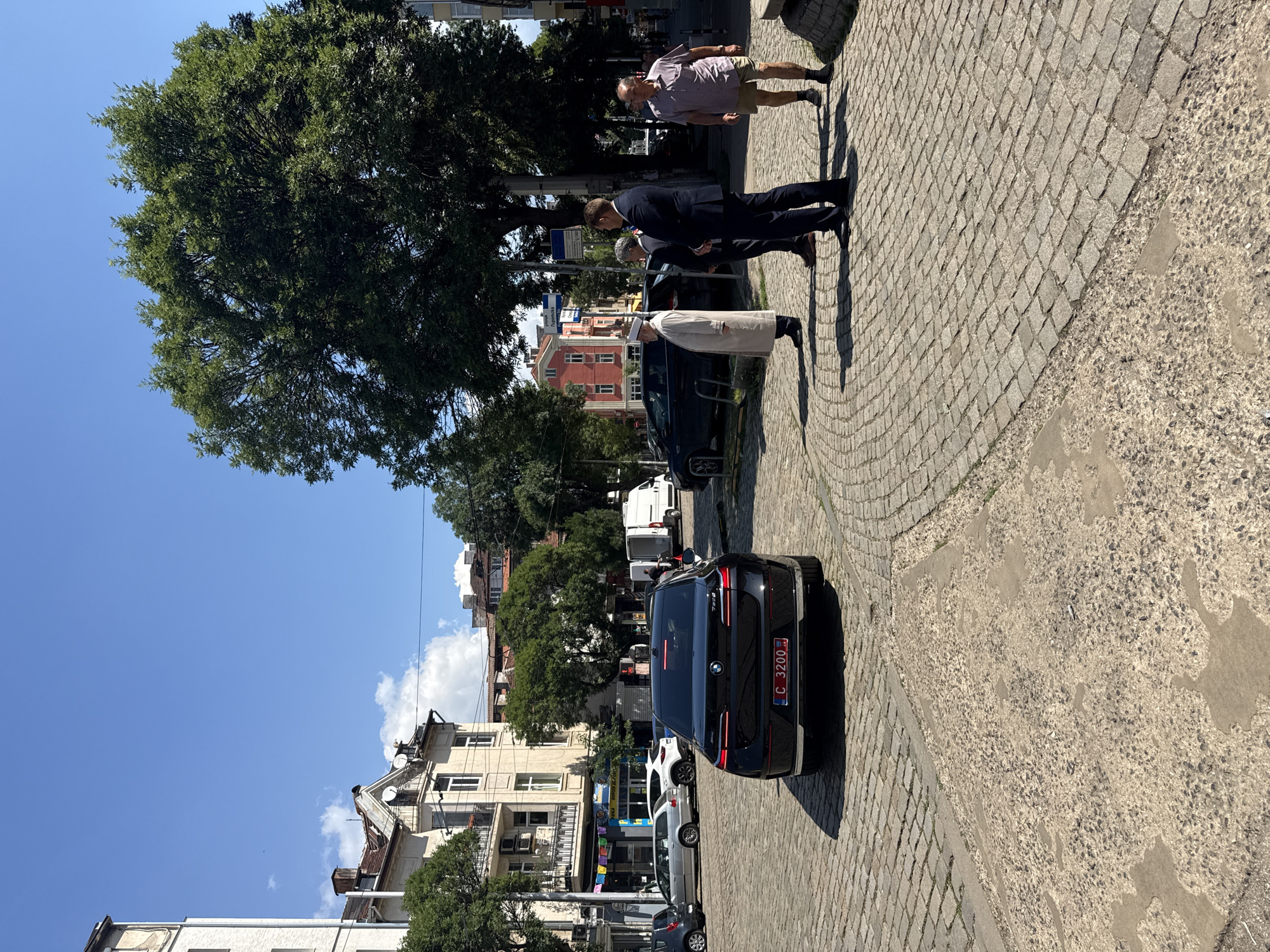 The image captures a street scene in Sofia, Bulgaria, on a sunny day. In the foreground, a black BMW i7 sedan is parked on a cobblestone street, its license plate reading "C 3200 B G". Several people are gathered on the cobblestone pavement to the right of the car. Three men are standing in a group; one in a dark suit, one in a lighter suit, and another in a light-colored robe and white hat. Further to the right, an older man in a short-sleeved shirt and shorts stands looking away from the camera. In the background, there are several buildings, including a multi-story building with balconies and an apartment building with a red facade. Trees line the street, and the sky is clear blue with a few wispy clouds. A white van is also visible in the background, along with other parked cars. The environment suggests a typical urban setting in a European city.