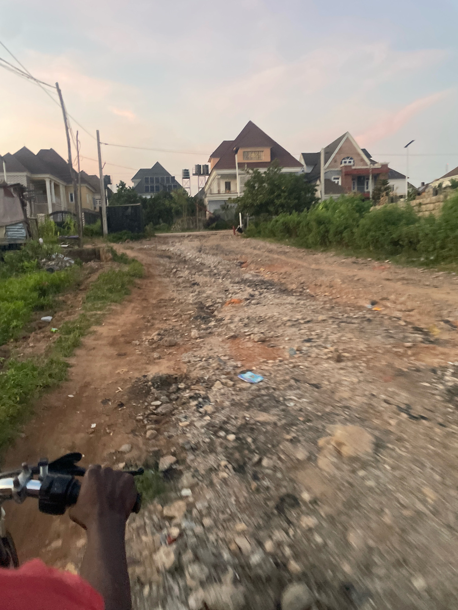 This is a medium-shot, ground-level, slightly angled, vertical photograph taken from the perspective of someone riding a motorcycle on a dirt road in Abuja, Nigeria. The sky is pale and overcast with hints of pink, suggesting either dawn or dusk. The road is unpaved and rough, with visible rocks and debris, indicating it is likely a developing area of the city.
On the left side of the road, there is overgrown green vegetation and some scattered trash. Beyond the vegetation, several modern-looking houses with pitched roofs and multiple stories are visible. These houses appear to be recently constructed or under construction. Some have balconies and decorative architectural features. There are also utility poles and electrical wires crisscrossing the sky.
On the right side of the road, the terrain is also rough and uneven, with more rocks and sparse vegetation. The road continues into the distance, flanked by more greenery and houses. The motion blur suggests the motorcycle is moving at a moderate speed. In the foreground, a person's hand, appearing to be dark-skinned, is gripping the handlebars of the motorcycle. The person is wearing a red shirt. The overall impression is of a suburban or semi-urban area of Abuja undergoing development, with a mix of modern housing and unkempt infrastructure.
