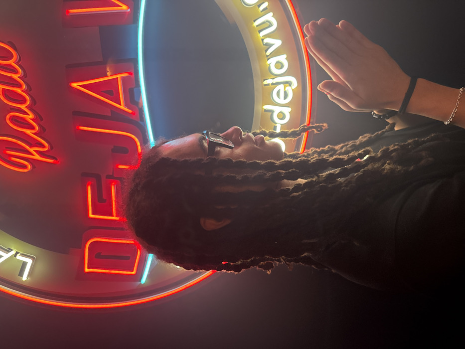 A person with long dreadlocks and glasses poses with their hands pressed together, palms facing each other. The background includes a round, neon sign featuring the word "Radio" in red, followed by "DEJAVU" in red and blue neon lettering. The sign also has additional neon accents. The lighting is dim, creating a dramatic effect and emphasizing the neon glow. The person is wearing a dark-colored shirt.