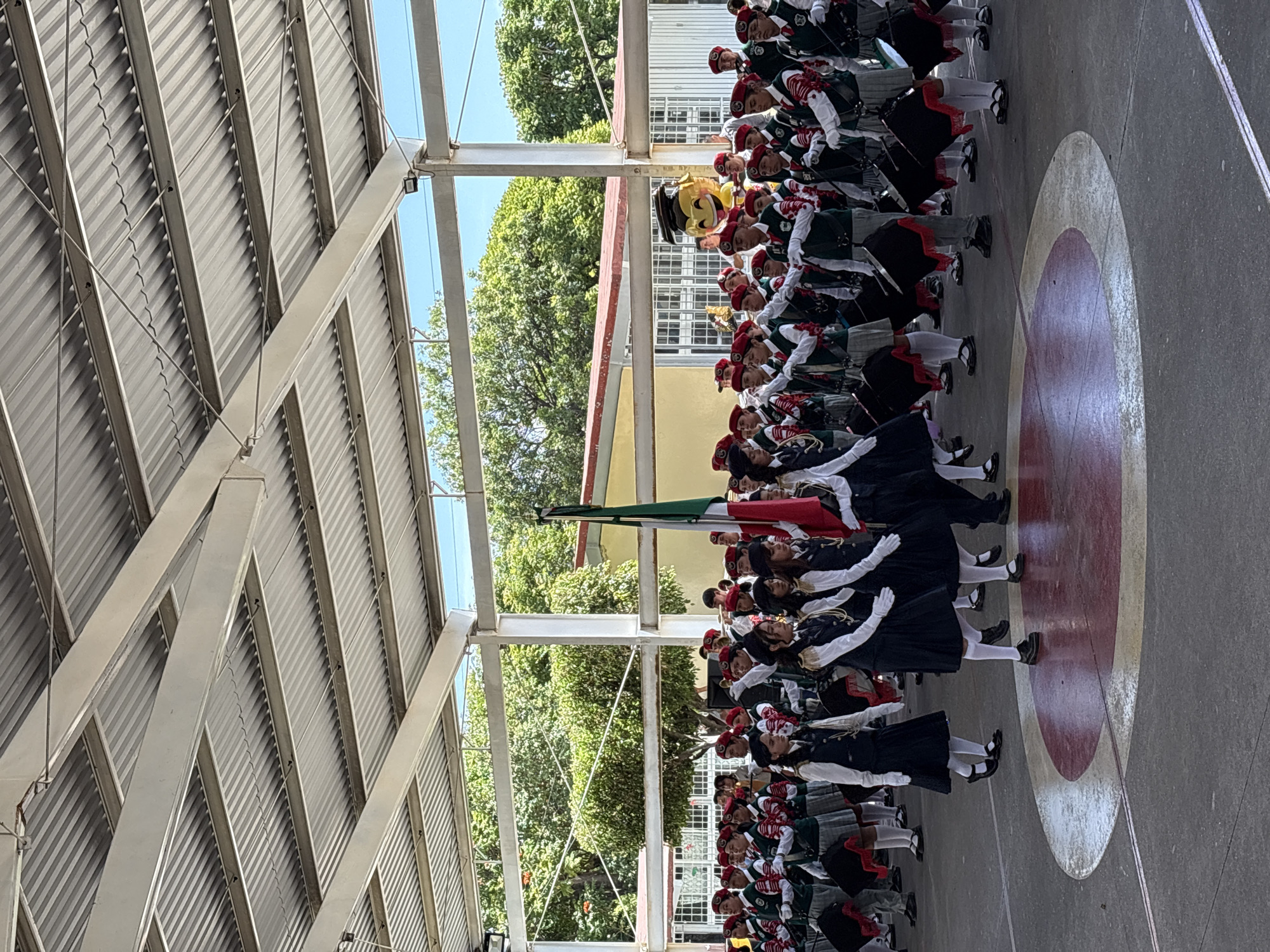 A marching band composed of young people in uniform is parading outdoors under a covered walkway. The band members are wearing dark skirts, white shirts, dark vests, and red berets. Many are carrying large drums. In the background, a mascot character with a graduation cap and a smiling face is visible. A Mexican flag is being held by one of the band members. The ground is a paved surface with a circular design. The location is identified as the city of Tejalpa, Mexico.