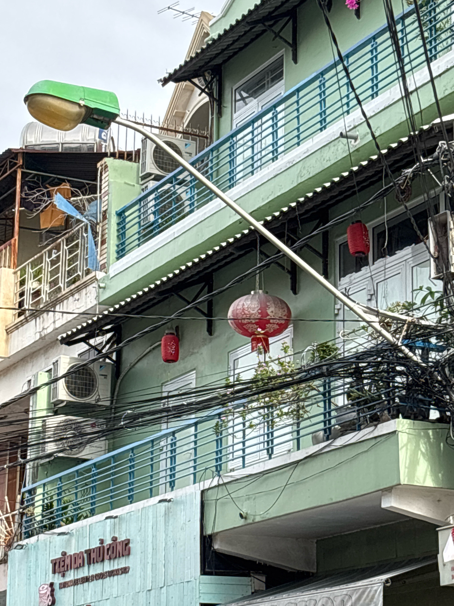 The image captures a close-up view of a light green building in Nha Trang, Vietnam. A street lamp with a green housing and a yellow bulb is prominently displayed on the left side. The building features multiple levels with blue balconies and white railings. Several air conditioning units are mounted on the exterior walls. Red lanterns are hanging from wires, adding a touch of cultural decoration. A dense network of electrical wires crisscrosses the facade, indicating a busy urban environment. The lower part of the building has a light blue facade with Vietnamese text in brown, which reads "TIEM DA THU CONG" and "HANDMADE LEATHER GOODS WORKSHOP". There are no people visible in the frame. The sky is overcast, suggesting a cloudy day.
