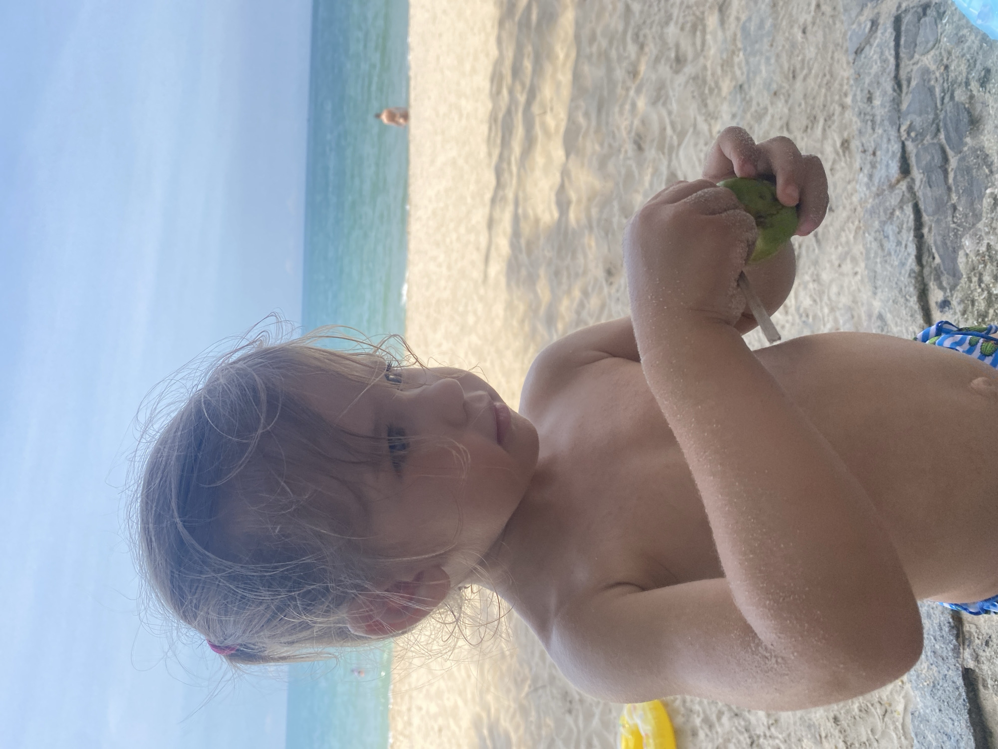A young girl, covered in sand, stands on a beach in Phu Thuy Ward, Vietnam. The girl is wearing blue and white patterned swim trunks and is holding a green object, possibly a lime or avocado, with a stick inserted into it. Her hair is light brown and pulled back with a pink hair tie. In the background, the ocean is calm and blue, with a person visible in the distance wading in the water. The beach is sandy, with shadows from unseen trees cast across the sand. A yellow inflatable ring is partially visible to the left of the girl.