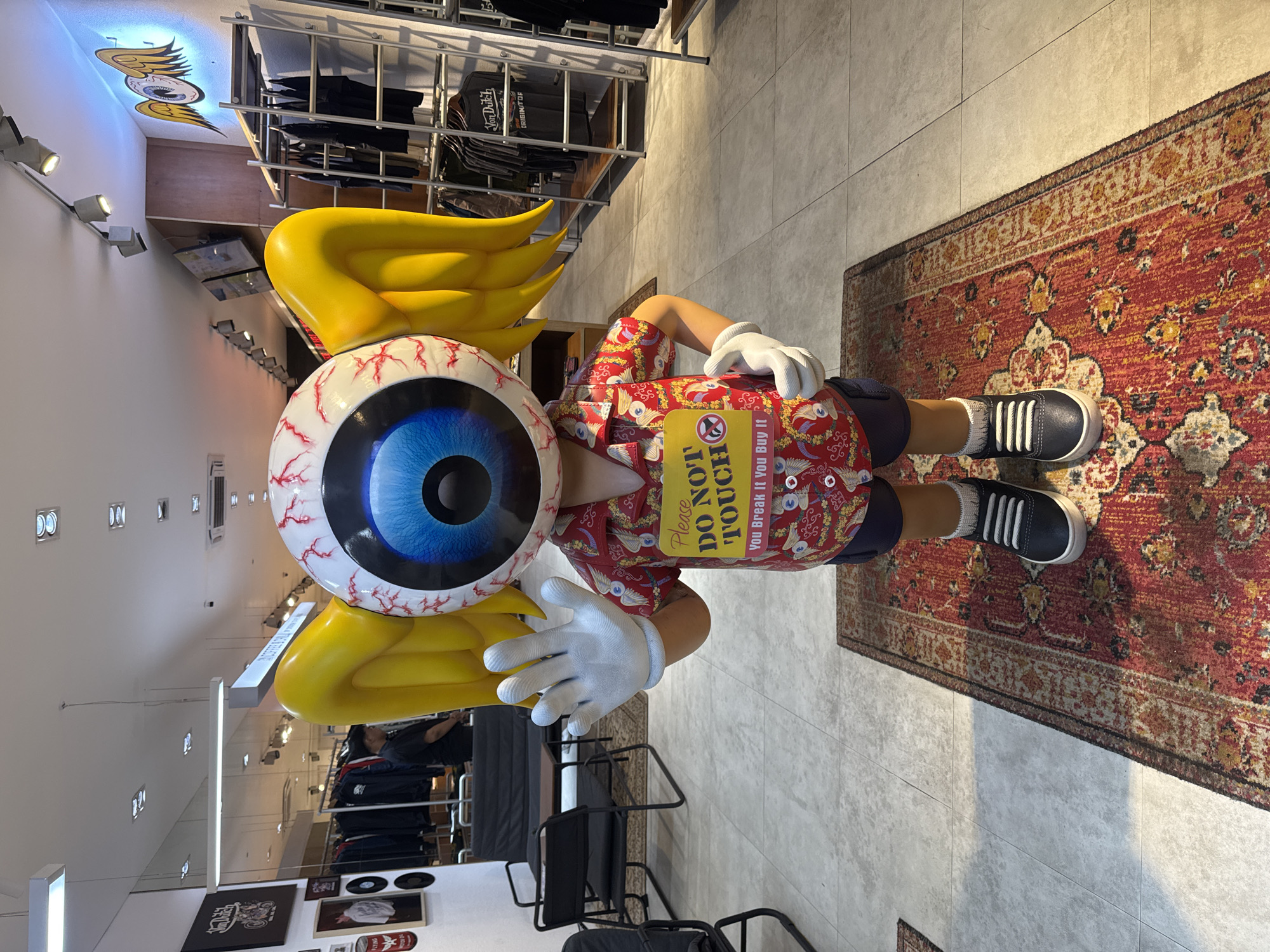 A life-sized, cartoonish statue of a figure with an oversized, realistic eyeball for a head stands centered in what appears to be a retail store. The statue wears a red, short-sleeved Hawaiian shirt with a white and yellow pattern, dark blue shorts, white gloves, and black sneakers with white laces and white socks. A yellow sign with red text reading "Please DO NOT TOUCH" and "You Break It You Buy It" is pinned to the statue's shirt. The statue is positioned on an oriental rug in the foreground, which is placed on a tiled floor. Behind the statue, rows of clothing racks filled with apparel are visible, along with a sofa and chairs in a seating area to the left. To the right, a metallic clothing rack displays various items, including t-shirts with branding. A mural of an eye with wings is painted on the wall behind the clothing racks. The overall environment suggests a trendy clothing boutique.