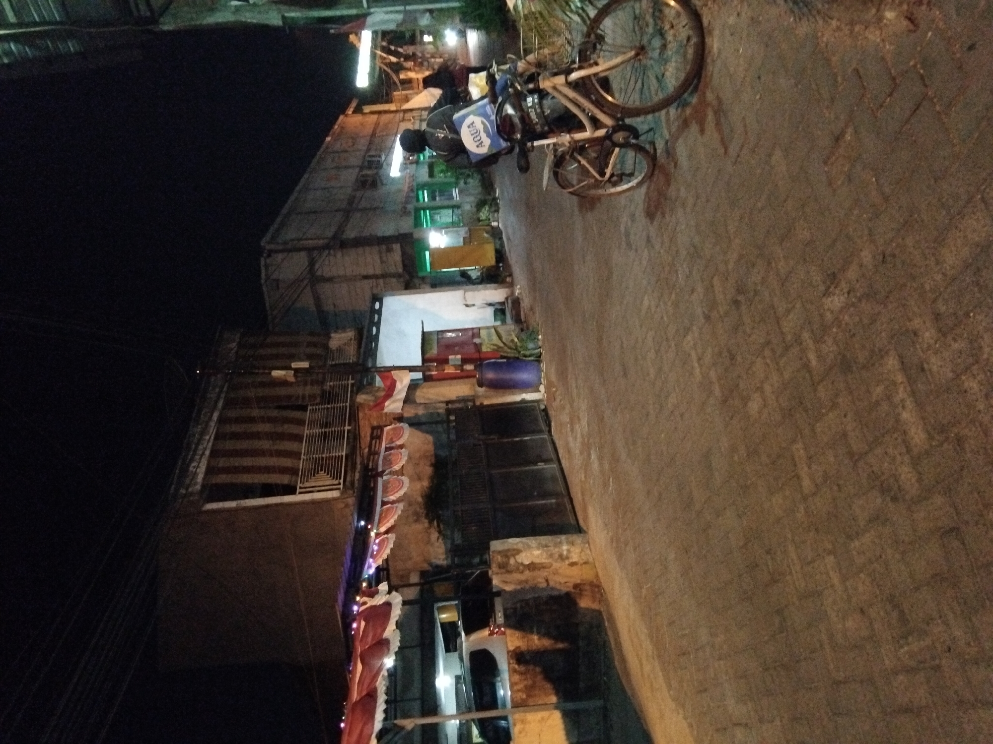 A street scene at night in Kepuh Kiriman, Indonesia, shows a paved walkway with a bicycle parked in the foreground. The bicycle has a basket in the front and a blue bag with the word "AQUA" on it strapped to the back. A person wearing a dark jacket and a black head covering is standing next to the bicycle, facing away from the camera. In the background, there are buildings with open doorways and windows, some illuminated with green light. A white car is visible parked behind a gate. Red and white banners with festive lights are draped across an awning. An Indonesian flag is visible on the right side of the frame. The ground is paved with interlocking bricks.