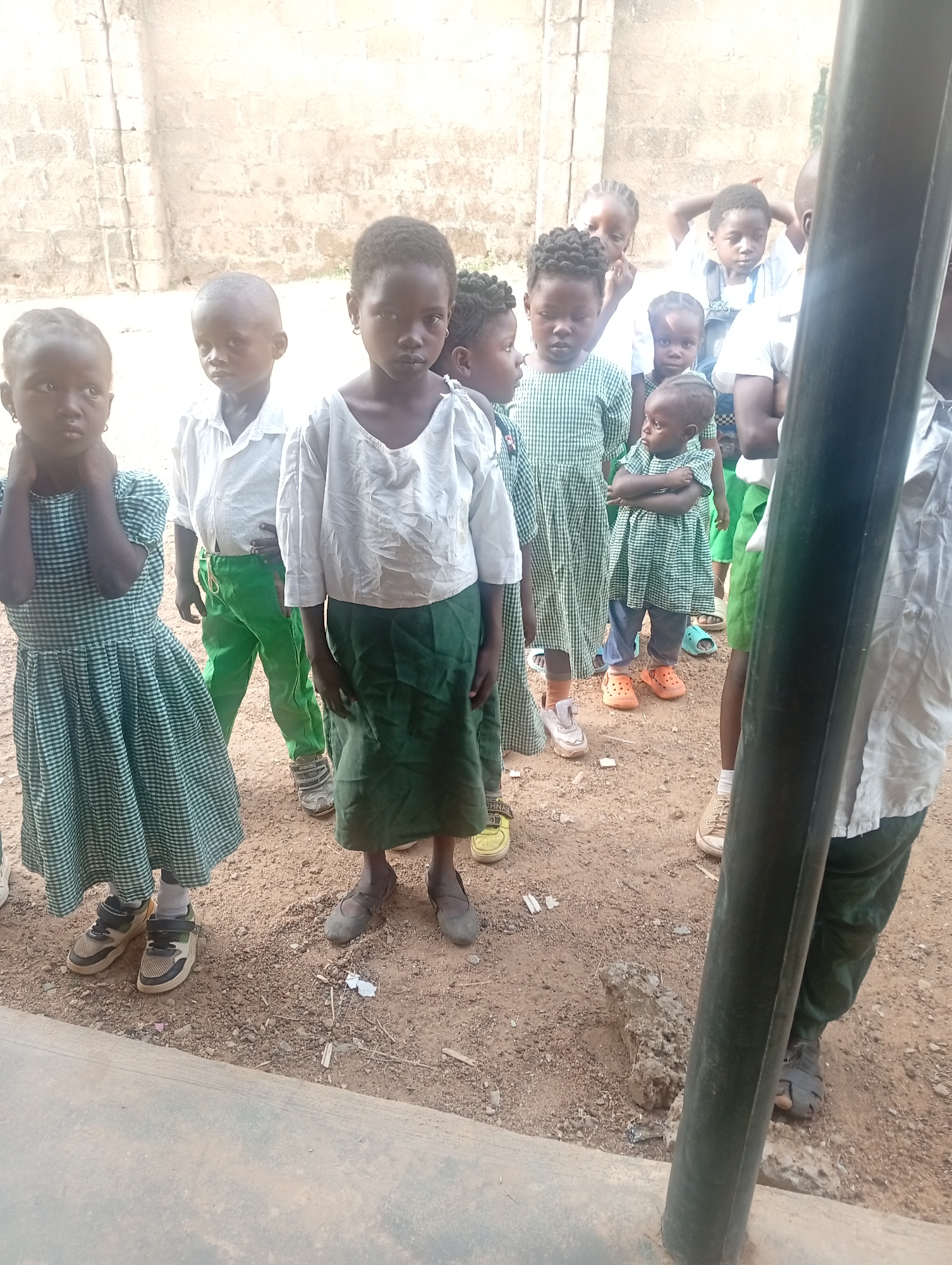 A group of schoolchildren are standing outdoors, likely in Narayi, Nigeria, given the request. They are primarily wearing uniforms consisting of white tops and green skirts or trousers. Some children wear green and white checked dresses. The children's footwear varies, including sneakers, sandals, and traditional shoes. The ground is dirt. A building wall is in the background. A large, dark pole is in the foreground, partially obscuring the view.