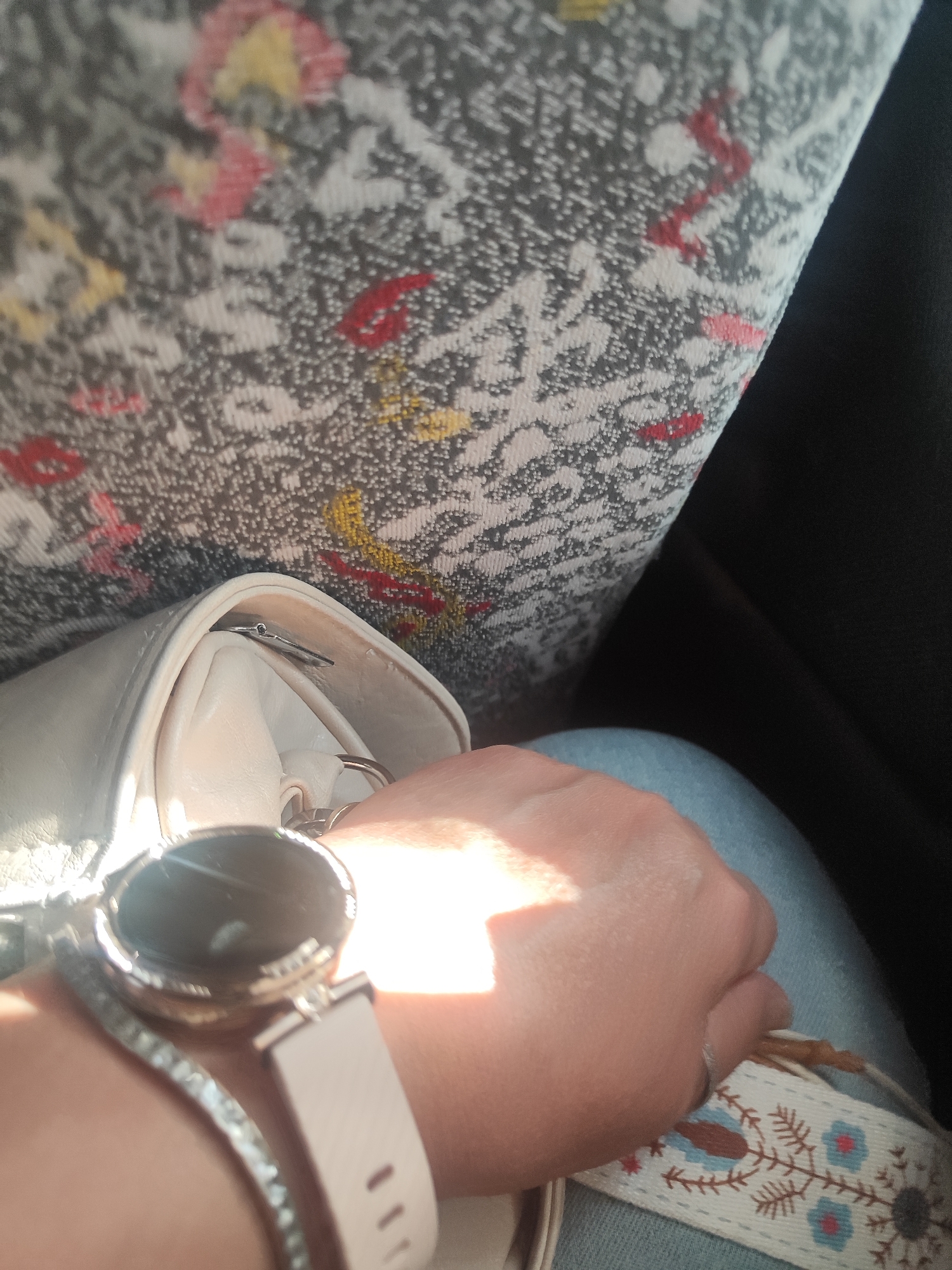 The image shows a close-up view of a person's left hand and arm, resting against a textured surface that appears to be a seat with a floral pattern in shades of gray, red, yellow, and pink. The person is wearing a light-colored smartwatch with a pink band and a decorative bracelet. The hand is illuminated by a strong light, suggesting it might be daytime, possibly with sunlight filtering through a window.
A portion of a light-colored purse is visible in the lower left quadrant, and a segment of blue denim fabric is seen in the lower right. A woven strap with a decorative embroidered pattern of blue flowers and brown branches is also visible around the person's wrist, partially covering the denim. The overall impression is one of casual relaxation, possibly during travel or a break. There is no visible text in the image. The lighting and the subject's relaxed pose suggest a calm, possibly indoor or shaded outdoor setting during daylight hours.