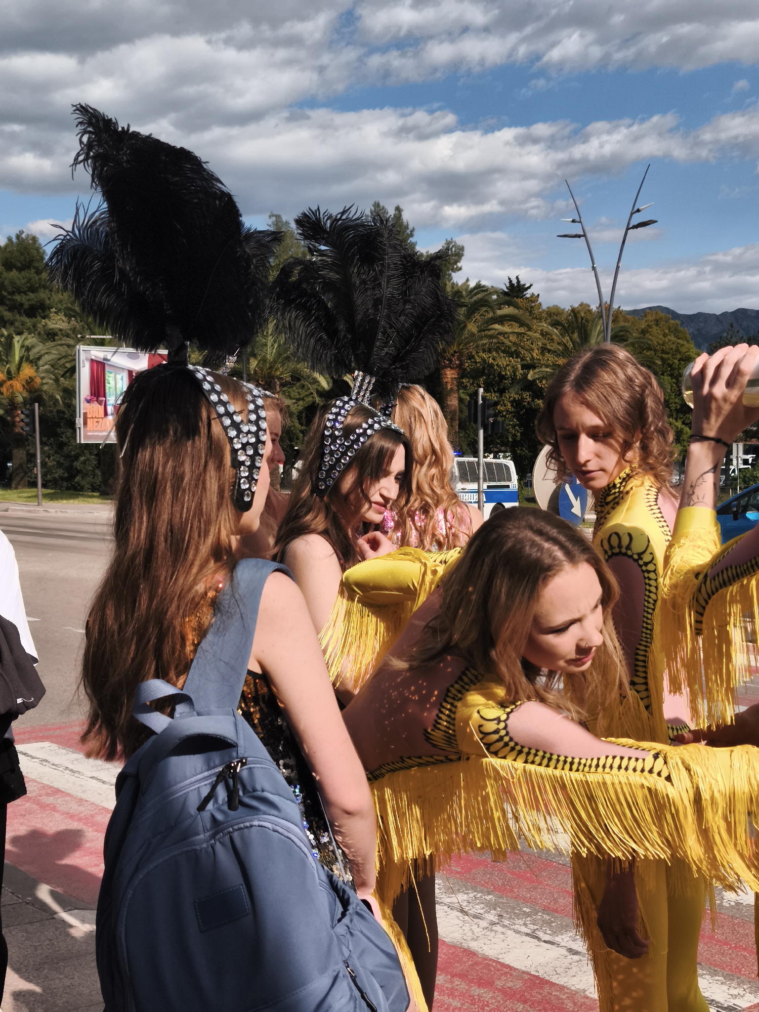A group of five individuals is gathered on a pedestrian crossing, likely interacting or posing for a photograph. Four of them are wearing elaborate costumes consisting of yellow fringed outfits, and black headdresses with large black feathers. The fifth person has a blue backpack and is wearing a top with sequins.
The background includes palm trees, street lights, a building with signage, a white vehicle, and a mountain range under a partly cloudy sky. The city and country are Unknown.