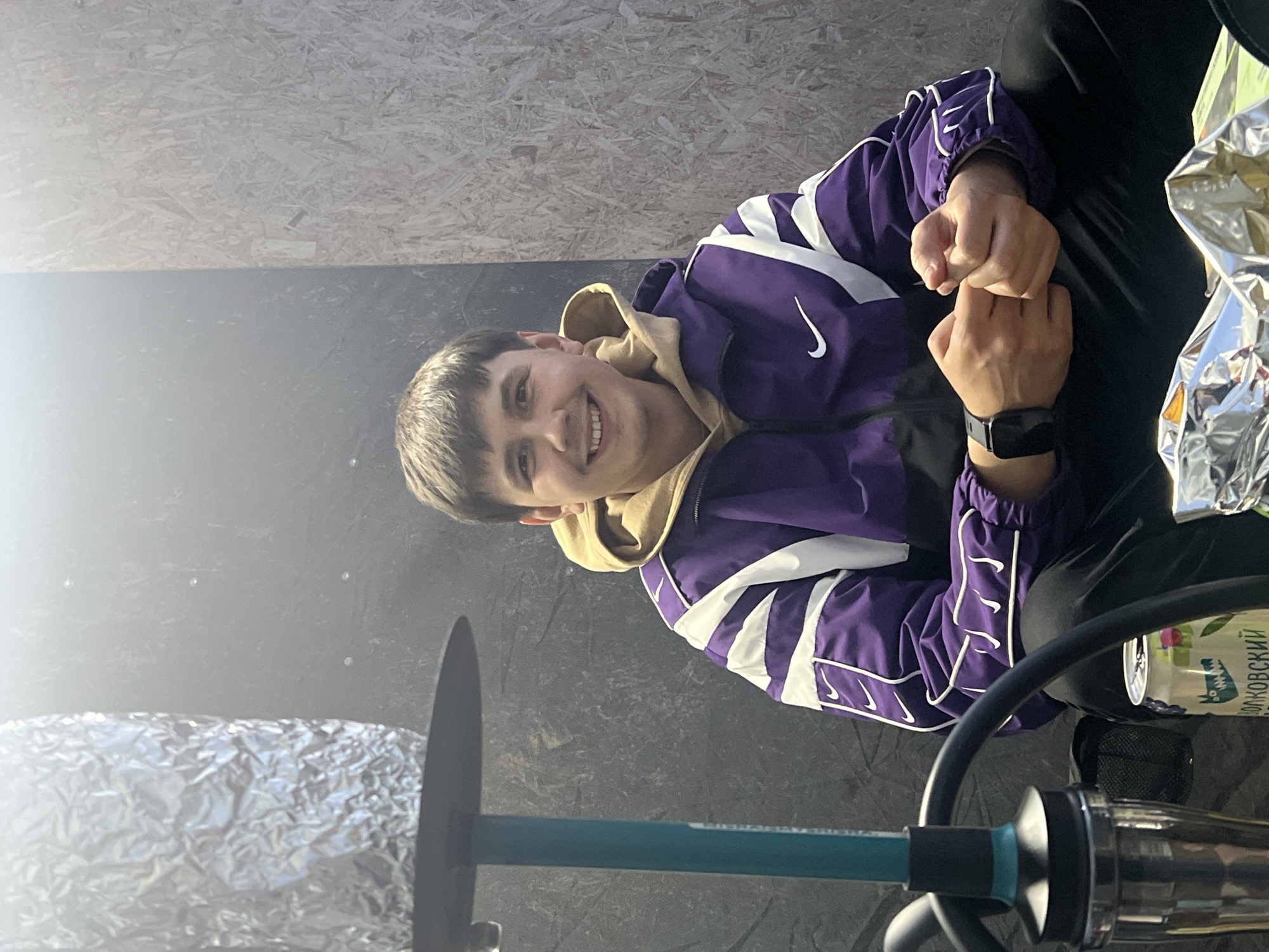 A young adult male is seated on a dark surface, smiling directly at the camera in an indoor setting in the city of Chaikovsky, Russia. He has short brown hair and is wearing a purple and black athletic-style jacket with white stripes and multiple white Nike swoosh logos on the sleeves and one large white swoosh on the chest. A light brown hooded sweatshirt is visible beneath the jacket. He is wearing black pants and a dark-colored smartwatch on his left wrist. His hands are clasped together in front of him. To the left, a hookah pipe with a teal-colored stem and a dark base is present; its bowl and charcoal area are covered with crumpled silver aluminum foil. On the dark surface near his right leg is an aluminum can with a light green label depicting a cartoon alligator and Russian text, "Волковскии" (Volkovsky). The background consists of two distinct wall sections: a dark grey, textured wall to the left and a lighter, textured oriented strand board (OSB) plywood wall to the right. A faint, diffuse haze is visible in the upper left portion of the scene.