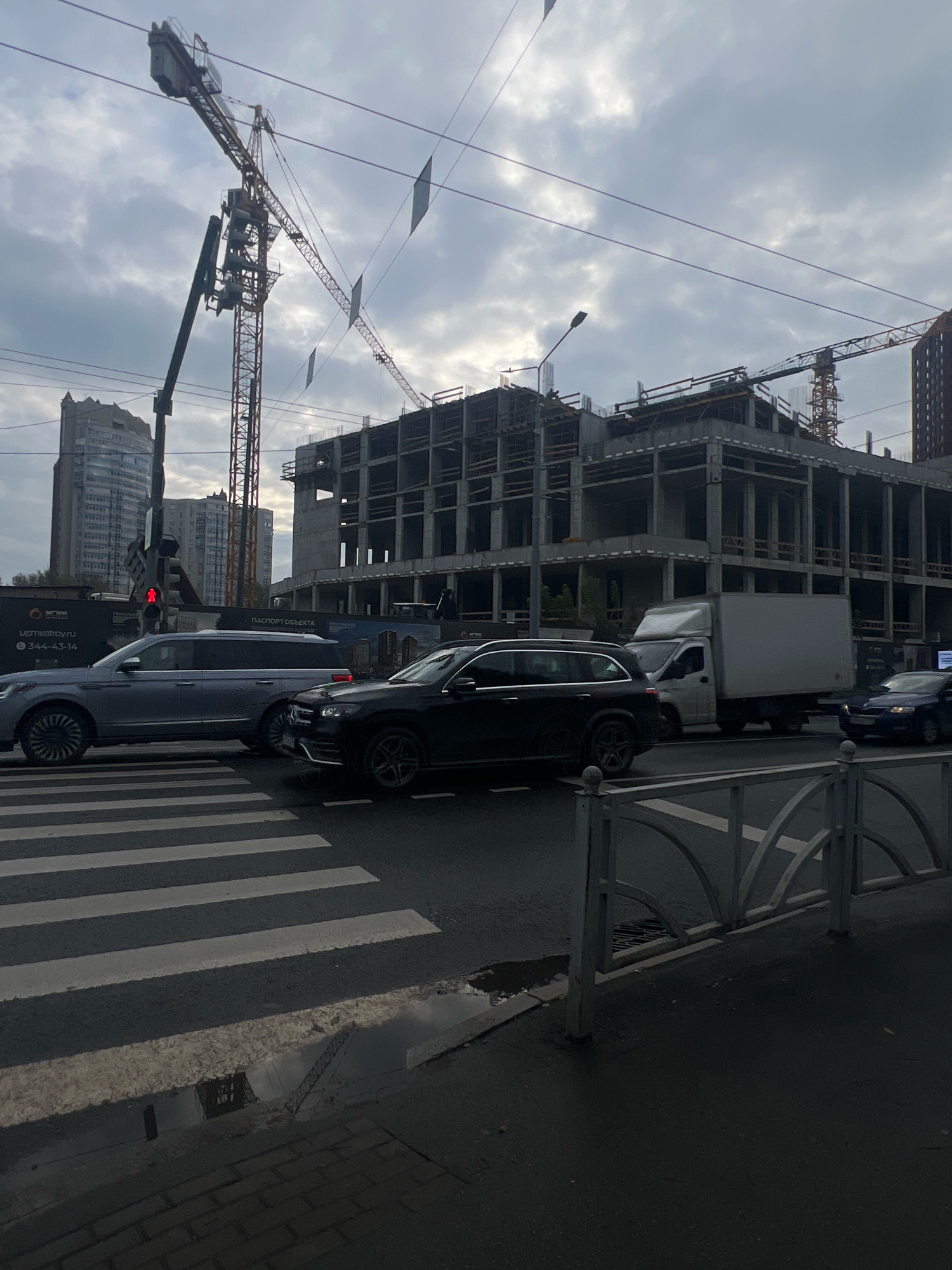 An overcast day in Yekaterinburg, Russia, where a construction site is visible, dominated by a large concrete building structure and multiple cranes. In the foreground, a pedestrian crossing is marked on a wet asphalt road. Several vehicles are present, including a silver SUV, a black SUV, a white box truck, and a dark-colored sedan. Traffic lights are visible, with one showing a red light. The scene appears to be a typical urban street with ongoing construction.