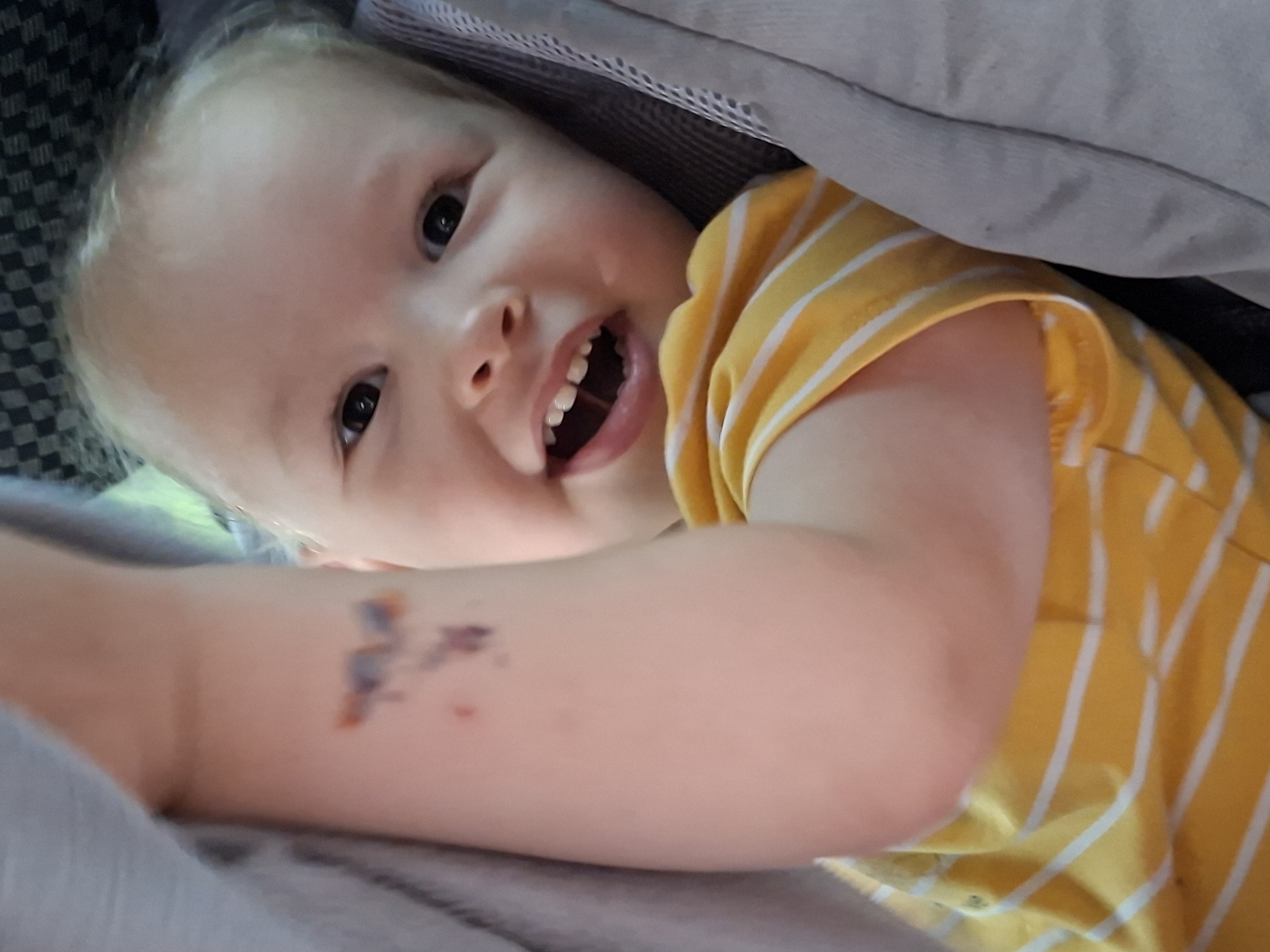The image shows a close-up of a young child with blond hair and dark eyes, smiling with their mouth open. The child is wearing a yellow and white striped t-shirt and has a small colorful sticker or temporary tattoo on their upper left arm. The child is partially covered by a gray fabric, possibly a blanket or clothing. The background is blurred, but a checkered pattern is visible at the top. The image does not contain any information that would allow for localization in Votkinsk, Russia.