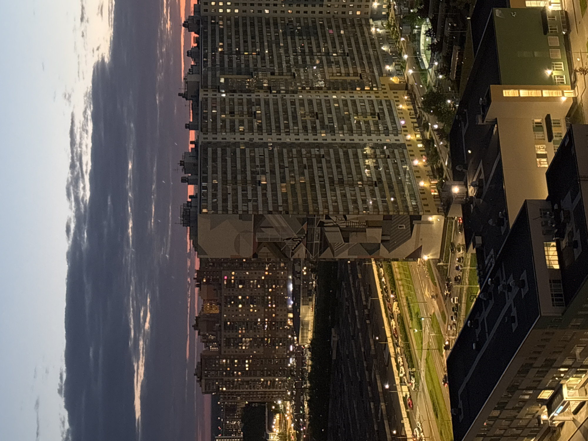 The image depicts an aerial view of Saint Petersburg, Russia, at dusk. Tall residential buildings with numerous illuminated windows dominate the landscape. A wide avenue with traffic is visible, lined with streetlights. Clouds fill the sky, with a hint of sunset colors near the horizon. Other buildings, some with green or yellow accents, are also visible.