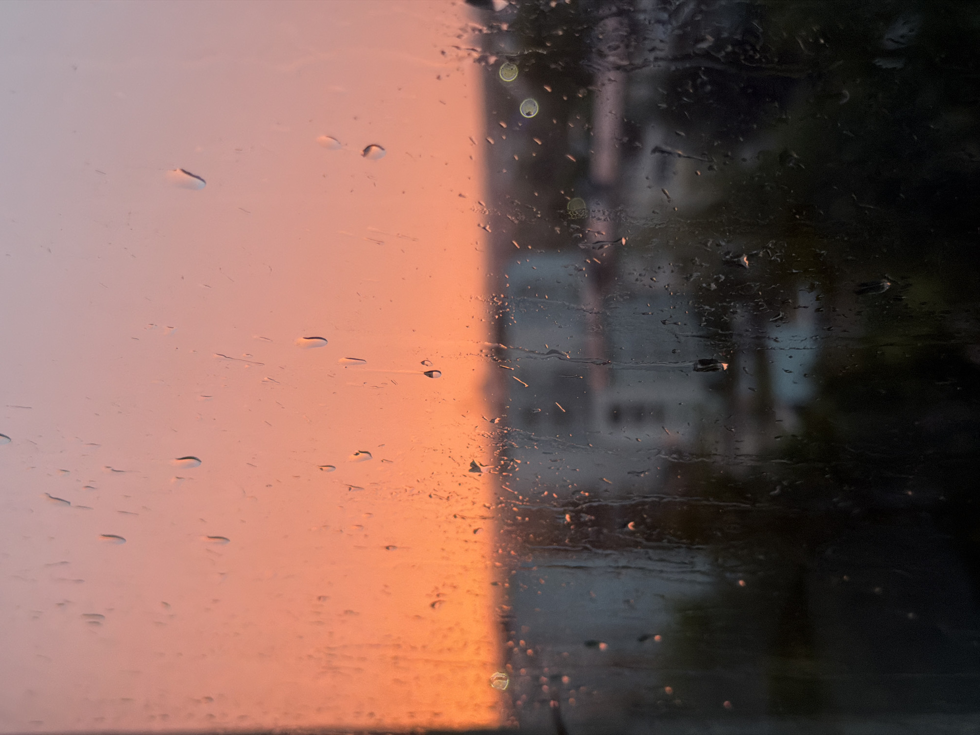 The image depicts a windowpane covered in raindrops, with a blurred cityscape visible beyond. The sky is a gradient of sunset colors, ranging from a vibrant orange at the horizon to a softer pink and pale blue higher up. In the mid-ground, indistinct buildings and trees are visible, softened by the rain and distance. The lower portion of the image is darker, showing more of the out-of-focus urban environment. The overall mood is atmospheric and serene, suggesting a moment captured during or after a rain shower at dusk in Chaikovsky, Russia.