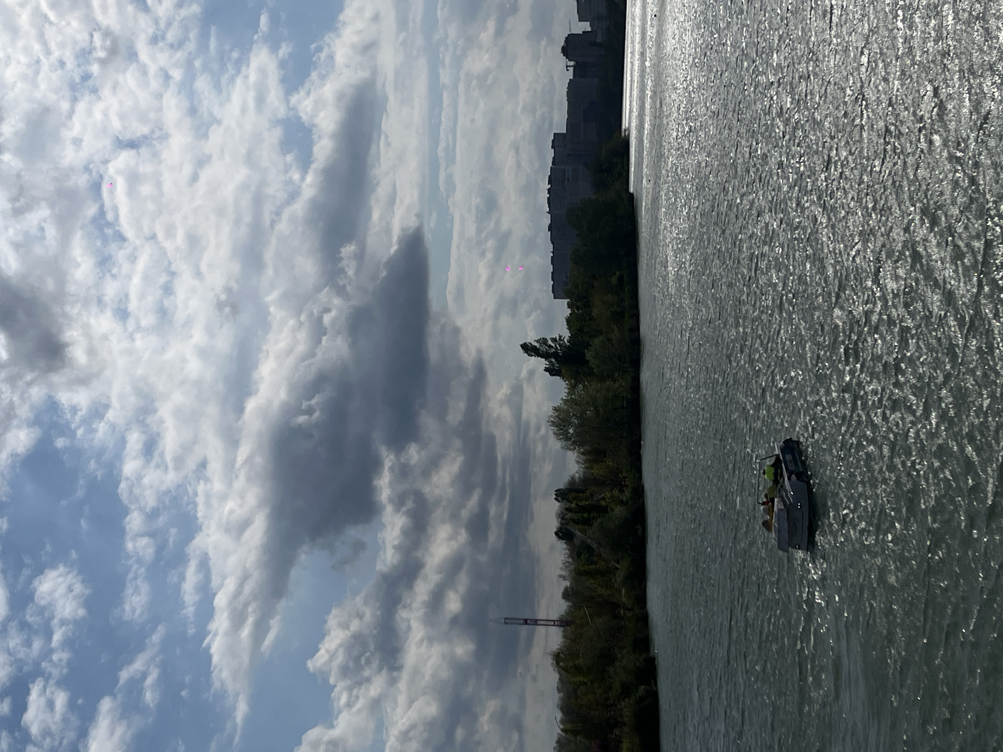 The image shows a view of the Kuban River in Krasnodar, Russia. Along the riverbank, there is dense vegetation with trees and bushes. In the distance, buildings can be seen. A small motorboat with people on board is traveling on the river. In the sky, there are numerous clouds. In the lower left is a thin cylindrical structure, possibly a tower.