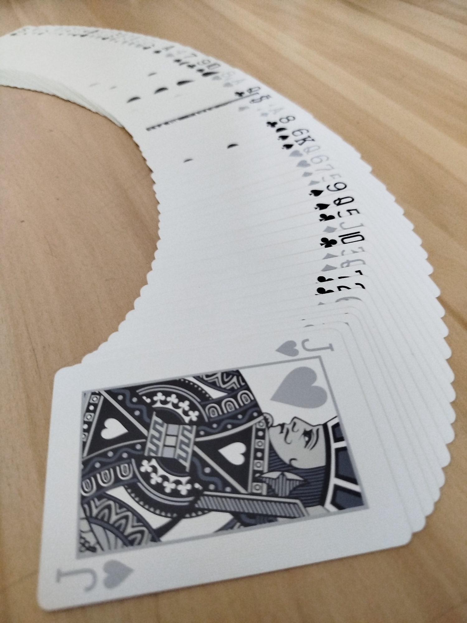 A deck of playing cards is fanned out on a light brown wooden surface. The cards are arranged in a curve, with the top of the deck visible and the bottom card fully in view. The bottom card is the Jack of Hearts, depicted in grayscale with a patterned background. The suits visible on the fanned cards include spades, clubs, diamonds, and hearts. The cards are positioned on a table, likely in an indoor setting. There is no indication of the city of Varna, Bulgaria, within the image.