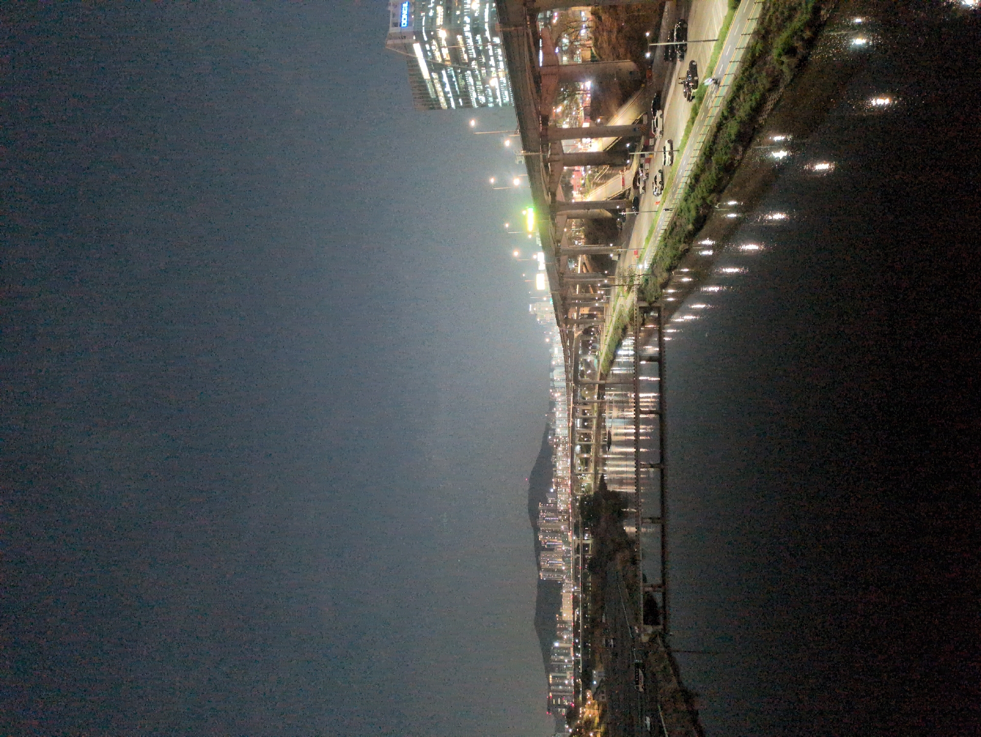 The image shows a night scene in Seoul, South Korea, featuring the Han River. A major highway, likely part of the Olympic Expressway, runs along the riverbank with multiple elevated sections and supporting pillars visible. Car lights and street lamps illuminate the highway and its surrounding area. The river reflects the city lights, creating a shimmering effect. A tall building, potentially the Dongil Tower, stands on the city side of the highway. The sky is dark, suggesting a clear night.