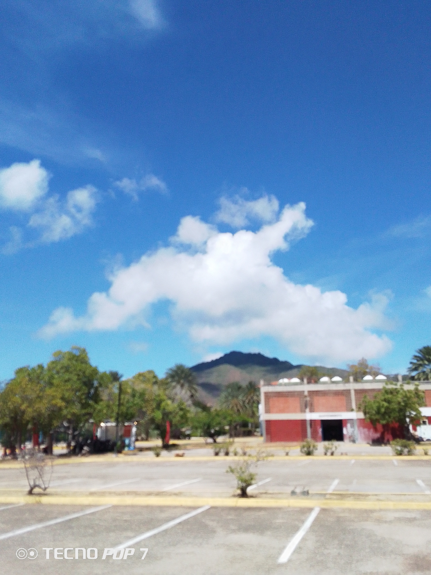 The image presents an exterior view of a building in Pedro González, Venezuela. The scene is dominated by a bright blue sky with scattered white clouds. In the background, a mountain is visible behind the building. The building itself is two stories tall, with a red and white exterior, featuring a series of arched windows on the top level. Trees surround the building, adding greenery to the landscape. In the foreground is an empty parking lot with white painted lines. The bottom left corner of the image contains the text "TECNO POP 7".