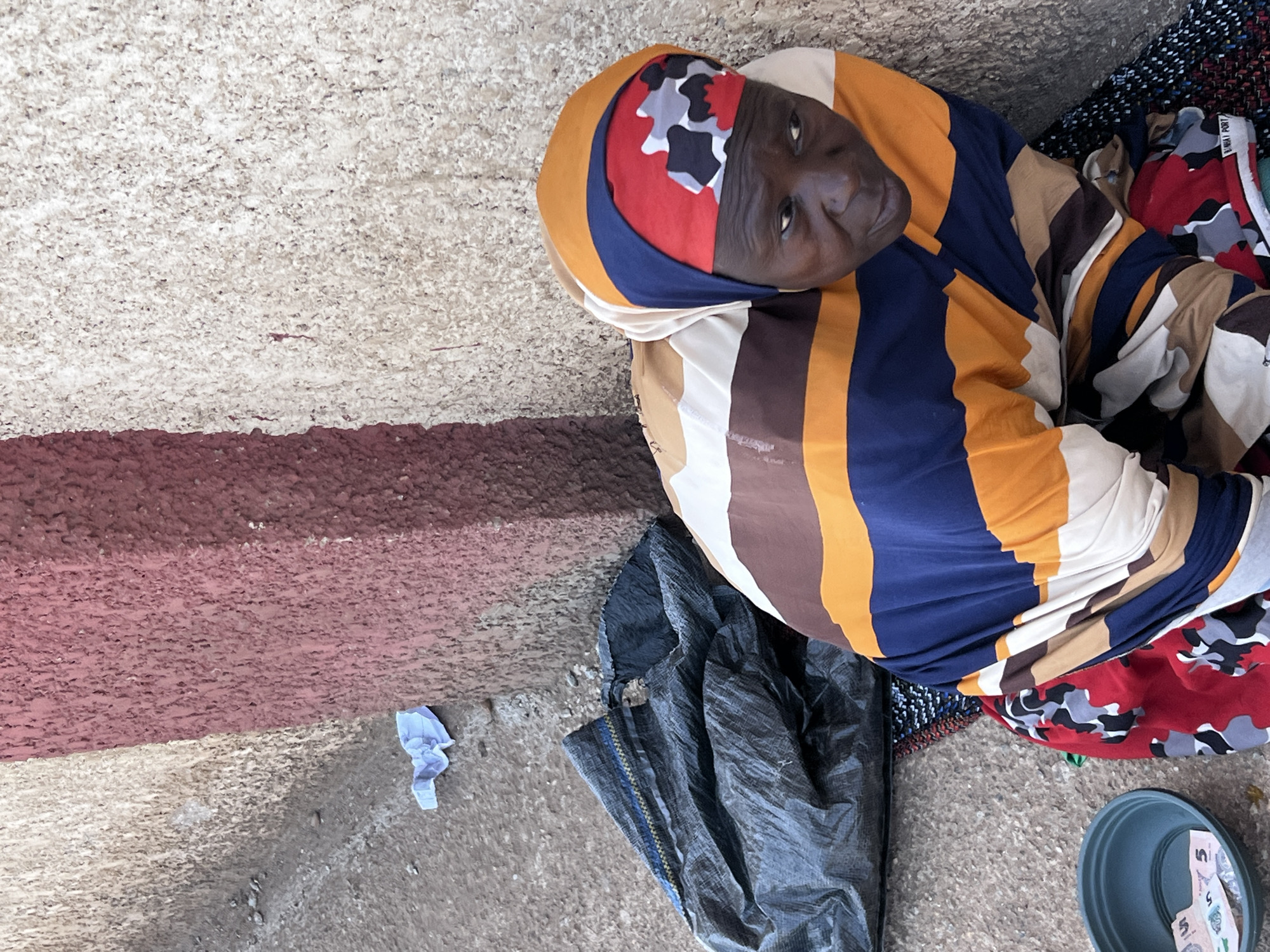 A photograph depicts an elderly woman on a concrete surface, presumably in an urban area in Jos, Nigeria. The woman, with dark skin and wrinkled features, is swathed in a multi-colored headscarf and striped clothing, her face visible as she looks upwards towards the camera. She rests on a layer of fabrics, including one with a camouflage pattern. Adjacent to her is a dark, possibly plastic, bag. A small blue plastic bowl containing currency notes sits to her lower right. The background shows a concrete structure, discolored in shades of red-brown and gray-white. A crumpled piece of blue paper is visible near the wall. The scene suggests a state of poverty and vulnerability in an urban setting.