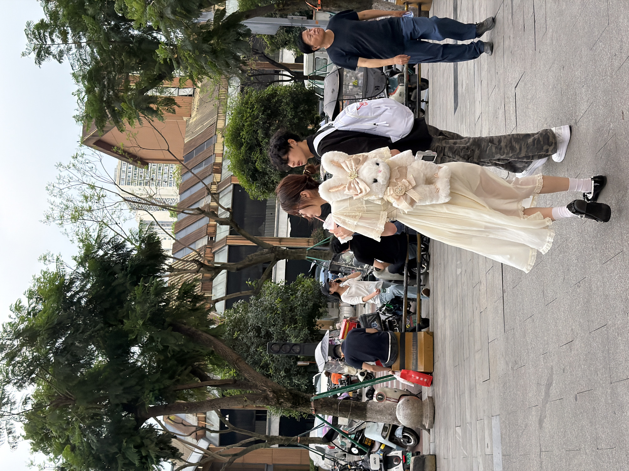 The image shows a street scene in Chongqing High-tech Zone, China. A young woman wearing a light-colored dress and a large plush bunny accessory stands next to a person with a white backpack. A man in a dark blue shirt and jeans stands nearby. Several parked scooters line the side of the street. Other people are present in the background. Trees and buildings are visible, including a unique building with a corrugated metal facade.