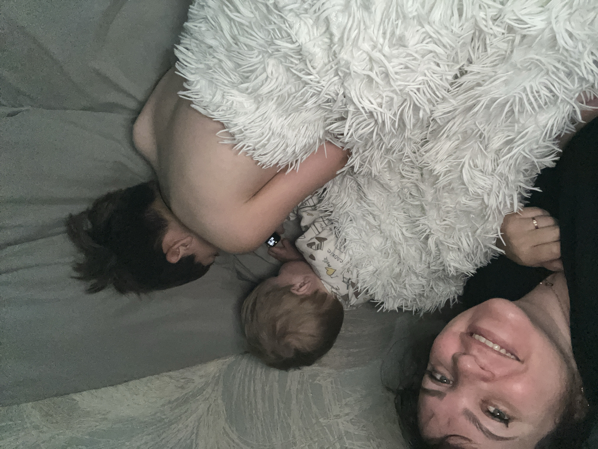 A woman, a child, and a person are lying on a bed. The woman is smiling at the camera. The child is looking down. The person is on their side, using a smartwatch. A white, furry blanket covers part of the scene. The location is unknown and cannot be identified as the city of Noviy, Russia.