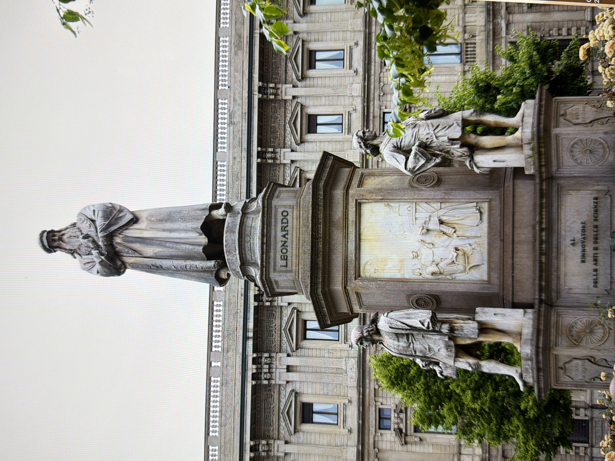 The image shows the Leonardo da Vinci Monument in Piazza della Scala, Milan, Italy. It is a grand monument featuring a central statue of Leonardo da Vinci standing tall, with four of his pupils at the base of the monument. The monument is made of bronze and is set against the backdrop of a large, ornate building. Trees and some flowering plants are visible in the foreground and on the sides of the monument. The sky is overcast, and the lighting is diffused, suggesting a cloudy day. The scene captures a well-known landmark in a public square.