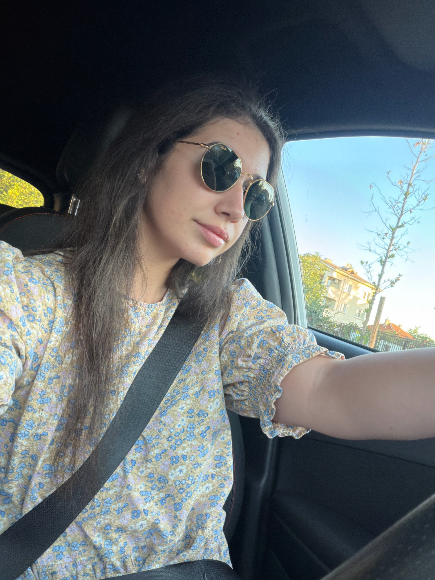 A medium shot captures a young woman with long, dark hair, wearing sunglasses and a floral print top, in the driver's seat of a car. She is looking away from the camera, towards the right side of the frame. A black seatbelt is visible across her chest. The interior of the car is dark, with the dashboard and steering wheel partially visible. Through the car window, a bright blue sky and trees are seen, along with buildings that appear to be in a residential area. The lighting suggests it is daytime, likely late afternoon. The presence of buildings with windows and balconies, along with the street layout, are consistent with urban residential architecture found in Sofia, Bulgaria.