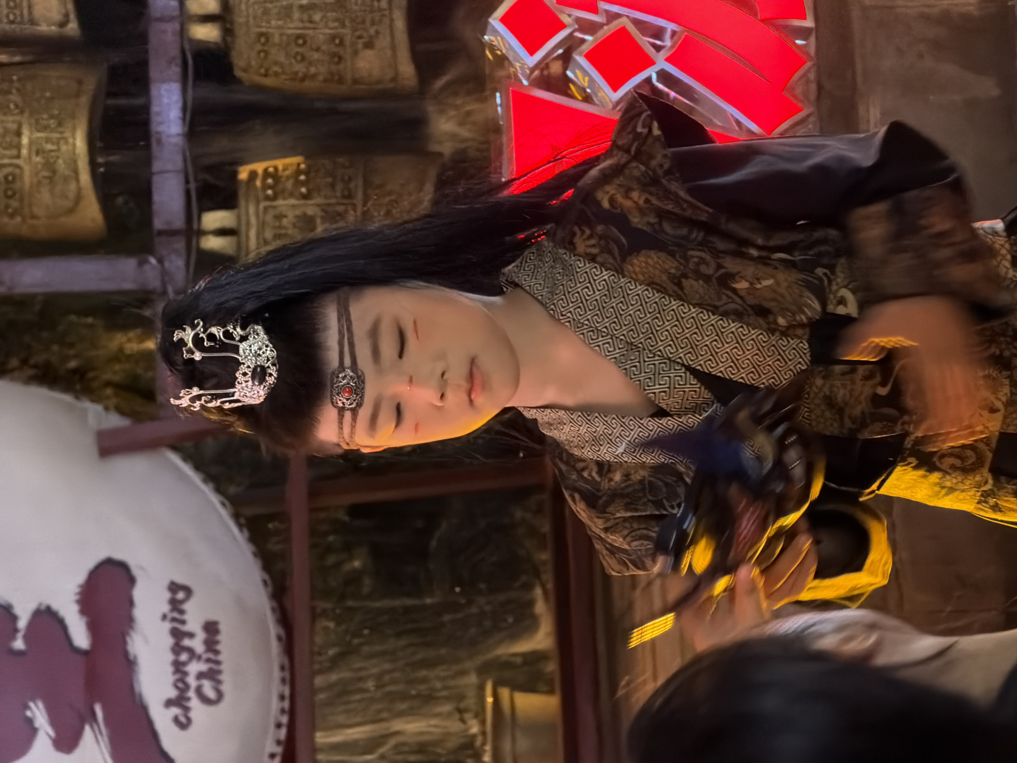 The image shows a young person in traditional Chinese costume, likely participating in a cultural performance in Yuzhong District, Chongqing, China. The person has their hair styled in a high ponytail and is wearing a silver headdress with black accents. Red markings are visible on their cheeks, and they are adorned with a decorative band across their forehead. Their attire consists of a dark outer garment with intricate patterns, layered over a lighter, patterned fabric. In their hands, they are holding a dark, mask-like object with blue and yellow accents. To the right of the person, a large, illuminated red geometric structure is visible, and in the background, a series of ornate, golden bells are displayed. The setting appears to be an indoor venue with warm lighting, possibly a theater or cultural exhibition space. The text "Chongqing China" is partially visible on a white circular banner in the upper left corner of the image.