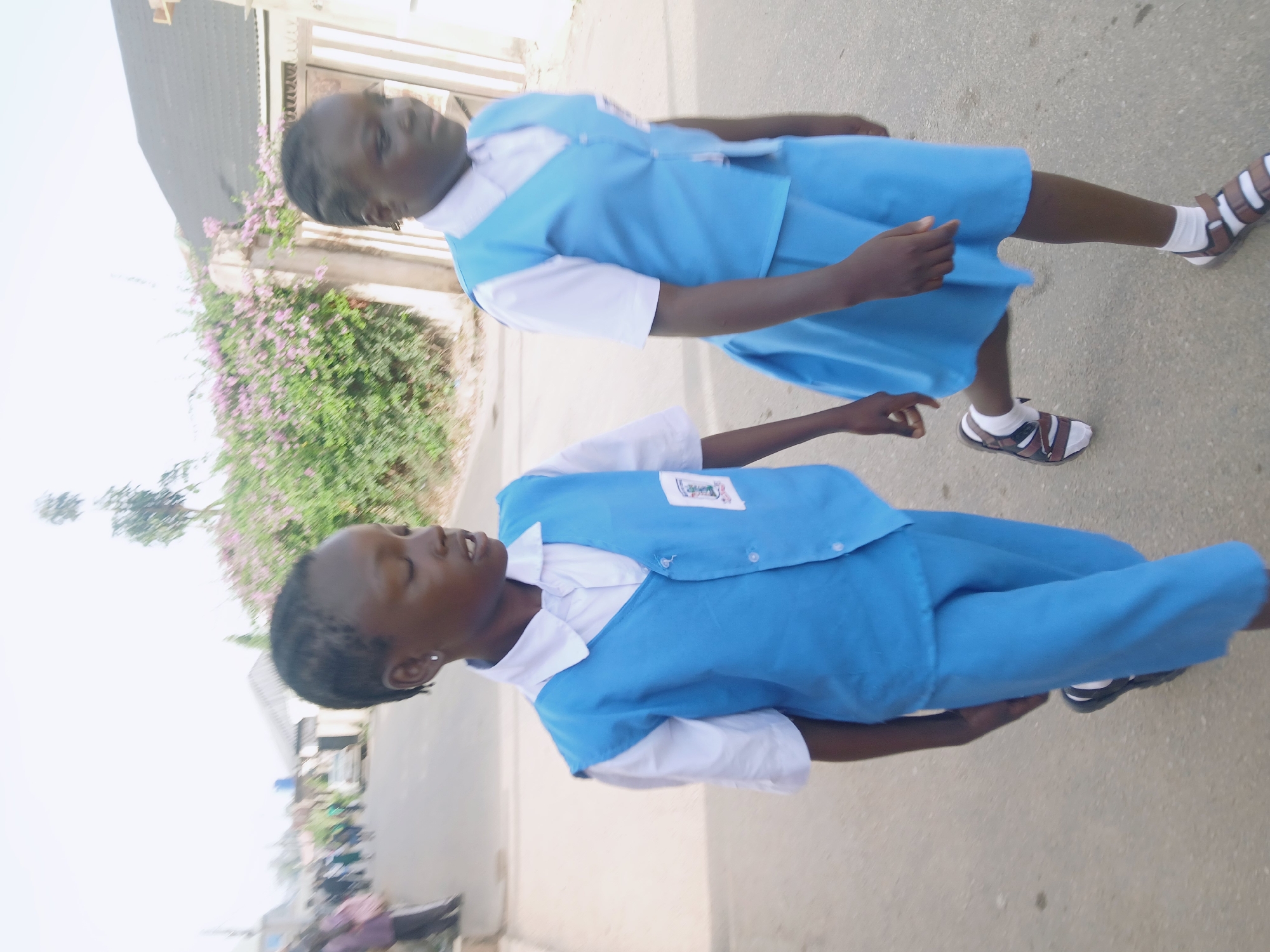 Two young girls, appearing to be school-aged, walk forward on a paved path or street during the day. The image is brightly lit, suggesting sunshine. Both girls have dark skin and short, dark braided hair.
In the foreground, a girl is walking with her body angled slightly towards the right. She wears a white, short-sleeved collared shirt beneath a light blue sleeveless vest, paired with matching light blue trousers. A small white patch or logo is visible on the left chest of her vest. Her eyes are partially closed, or cast slightly upwards. She wears white socks and dark-colored sandals.
Slightly behind and to the left, another girl is also walking, facing the same direction. She is dressed in a white, short-sleeved collared shirt underneath a light blue sleeveless dress. A similar white patch or logo can be seen on the left side of her dress. Her expression is neutral, looking forward. She wears white socks and brown sandals.
The background is softly blurred but reveals green foliage with scattered pink flowers on the left, and hints of buildings or structures on the right. The ground is a light gray, possibly asphalt or concrete. No discernible text is visible on the uniforms or in the background.