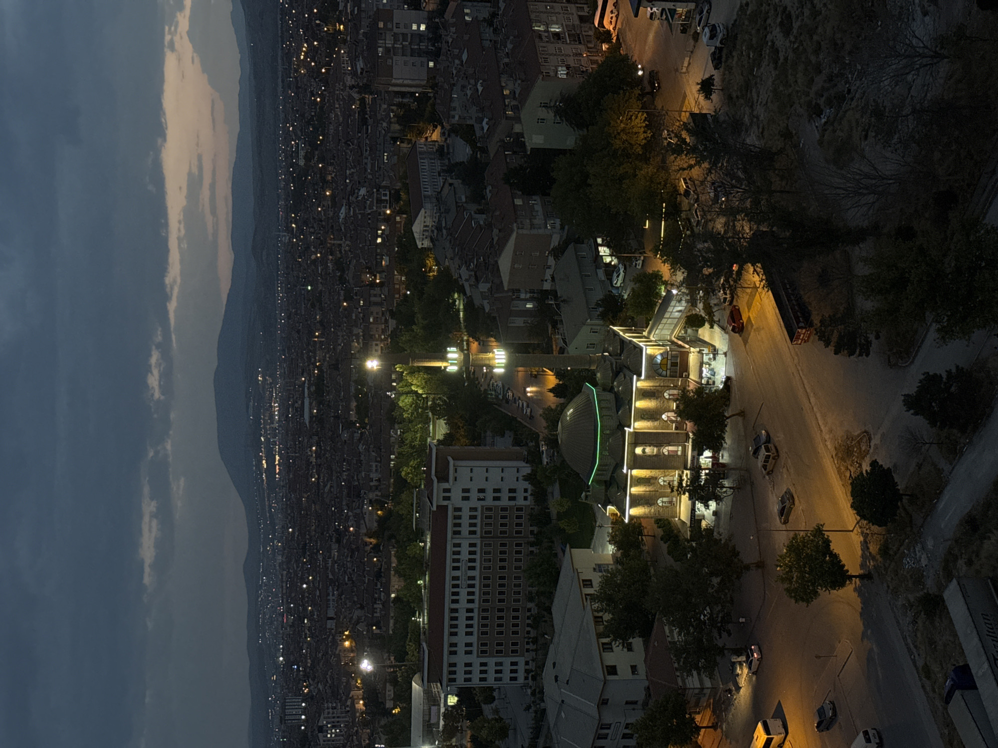 The aerial night-time view captures a segment of the city of Konya, Turkey. A prominent mosque with a green dome and illuminated minaret stands at the center, surrounded by various buildings, including a large, multi-story structure with many windows. Several cars are parked on the street and in parking areas around the mosque. In the background, the city extends into the distance with countless lights indicating a dense urban landscape. Distant hills form the horizon under a dark, cloudy sky. The scene suggests a peaceful evening in a developing urban area.