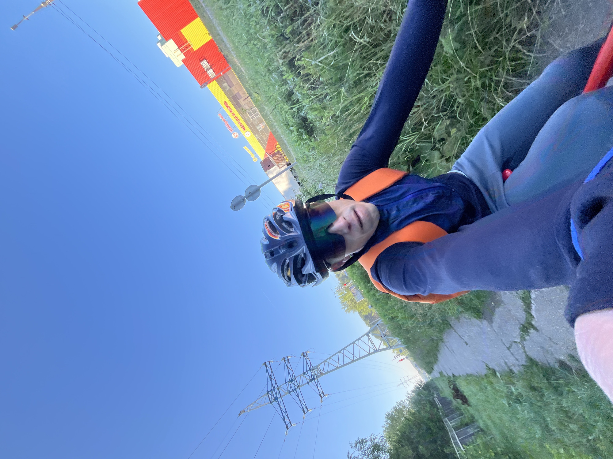 The image shows a first-person perspective of a person on a bicycle on a paved path in Chaikovsky, Russia. The person is wearing a helmet, sunglasses, a dark long-sleeved shirt, grey pants, and an orange vest. In the background, there is a large red and yellow building with Russian text, likely a commercial establishment. To the left, there is a tall power line pylon and lush green vegetation. The sky is clear and blue.