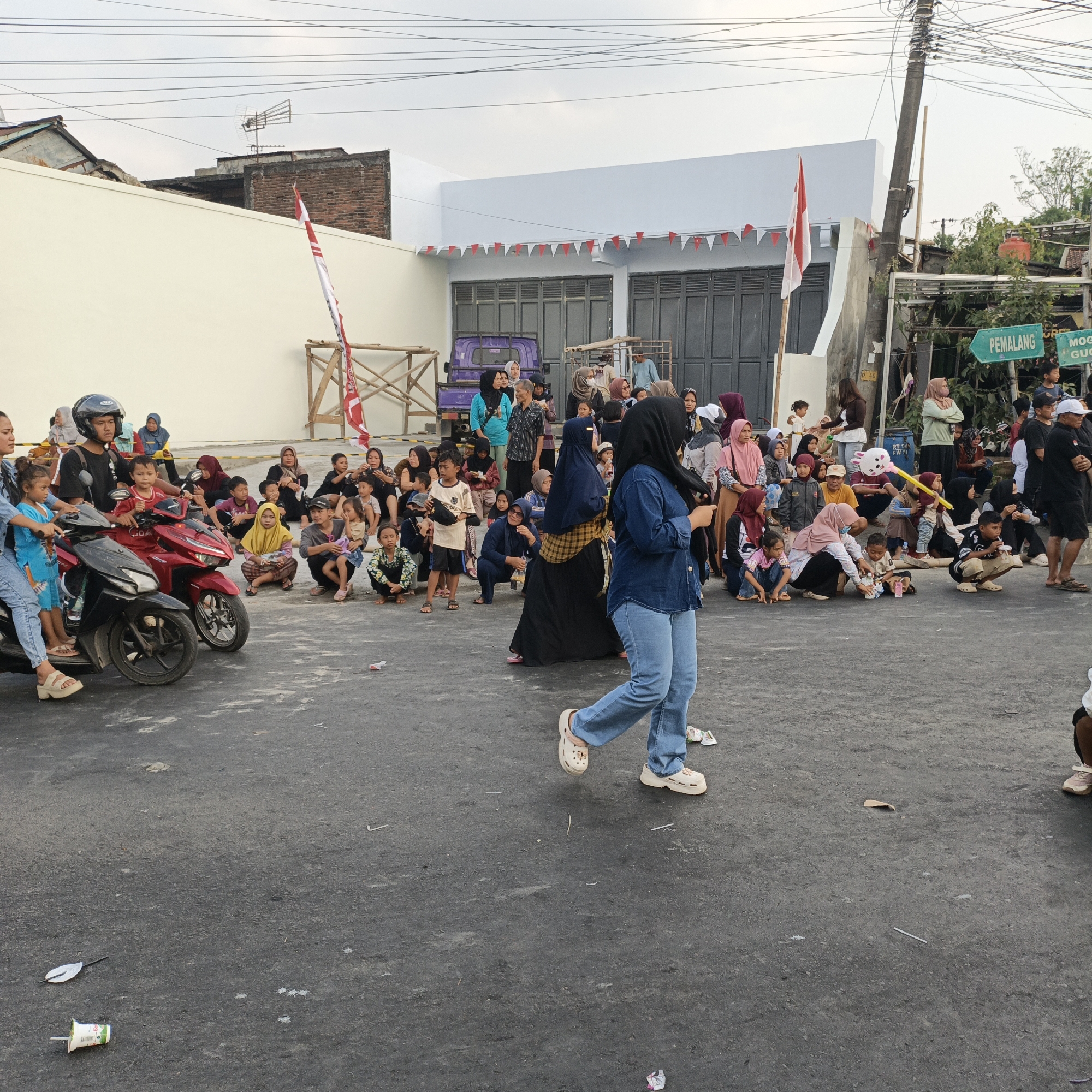 In Randudongkal, Indonesia, a crowd of approximately 60 people, predominantly women and children, are gathered on a paved road. The scene appears to be a community event, possibly a festival or celebration, indicated by the presence of Indonesian flags and red and white pennant banners strung across the front of a building. A group of individuals are seated on the ground in front of the building, while others stand or sit on motorcycles. Two motorcycles are parked on the left side of the image, with a woman and child seated on one. In the foreground, a woman in a blue denim jacket and jeans walks away from the camera. The building in the background is white with dark gray doors and windows. A sign in the upper right corner indicates directions to "PEMALANG" and "MOGE GUCU". The overall environment suggests an outdoor gathering in a residential or commercial area.