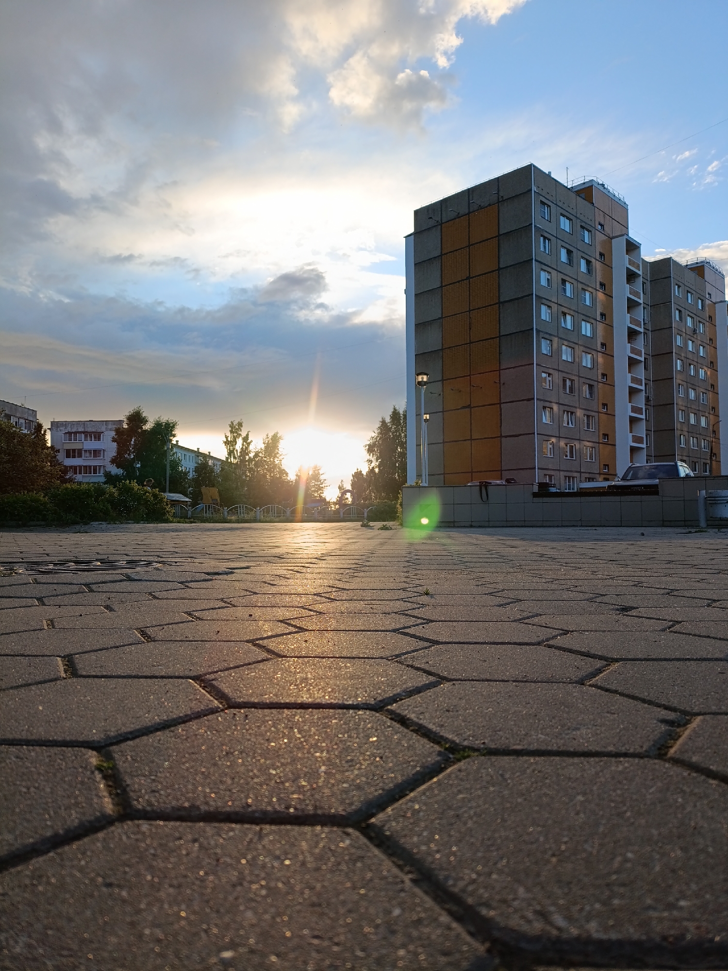 The image depicts a street-level view of a residential area in Chaikovsky, Russia, during sunset. The foreground is dominated by hexagonal paving stones, reflecting the warm, golden light of the setting sun. In the mid-ground, a row of trees separates the paved area from several apartment buildings. The buildings are multi-story structures with a facade of gray concrete and orange panels. One building on the right features a modern architectural style with a white columned entrance. A dark-colored car is parked in front of this building.
The sky is a dynamic mix of blue and dramatic clouds, with the sun intensely bright and low on the horizon, casting long shadows. The sunlight creates lens flare and illuminates the clouds with hues of yellow and orange. The overall mood is serene and peaceful, capturing the tranquil beauty of a late summer evening. The setting suggests a typical urban or suburban landscape in Russia. No text is visible in the image.