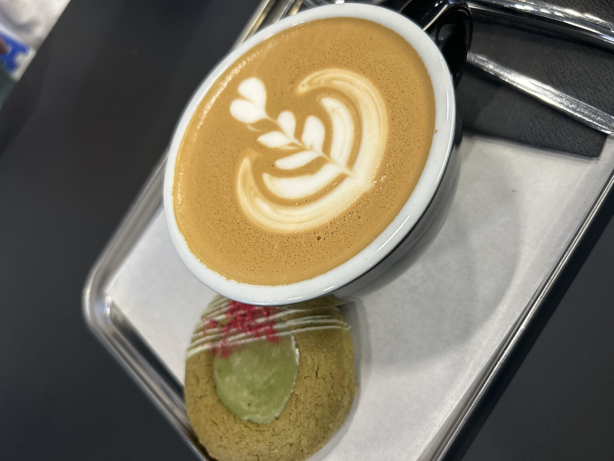 A latte with latte art is placed on a silver tray next to a cookie. The cookie has a green center and is drizzled with white icing and red sprinkles. The tray is on a black surface. There is silverware in the upper right corner of the image. The location of the image is not specified, though it is stated that the city is in Izhevsk, Russia. There is no other information about people, actions, or interactions.