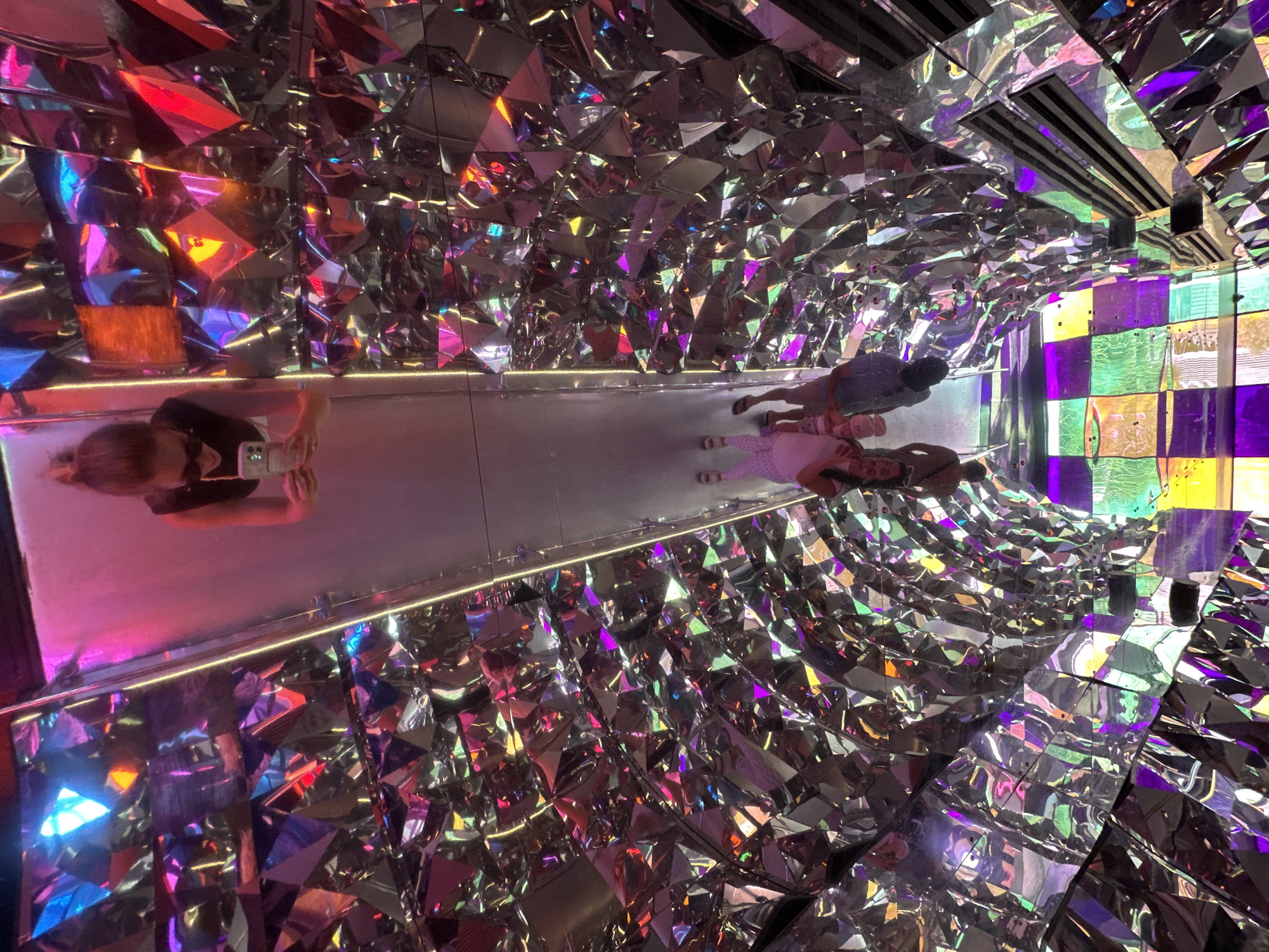 The image depicts an immersive art installation with a kaleidoscopic effect, likely located within the Weizmann Institute of Science in Rehovot, Israel, given the context of the original query which mentioned this location. The environment is characterized by a tunnel-like structure lined with highly reflective, faceted surfaces, creating a disorienting and vibrant visual experience. Bright colored lights, predominantly pink and blue, illuminate the space and are reflected infinitely by the mirrored surfaces.
In the upper portion of the image, a woman is seen from the chest up, taking a selfie with her phone. She is wearing sunglasses and has her hair pulled up. Her reflection is visible in the mirrored surfaces.
Further down the tunnel, also reflected in the mirrored walls, are two individuals. One is a woman wearing a white patterned dress and pink shoes. The other is a person wearing a blue shirt and dark pants. They appear to be walking or standing together.
The floor of the installation, visible towards the bottom of the image, is a grid of colored lights, predominantly green and purple, which further contributes to the psychedelic atmosphere. The overall impression is one of visual complexity and sensory stimulation, typical of an interactive light and mirror exhibition.