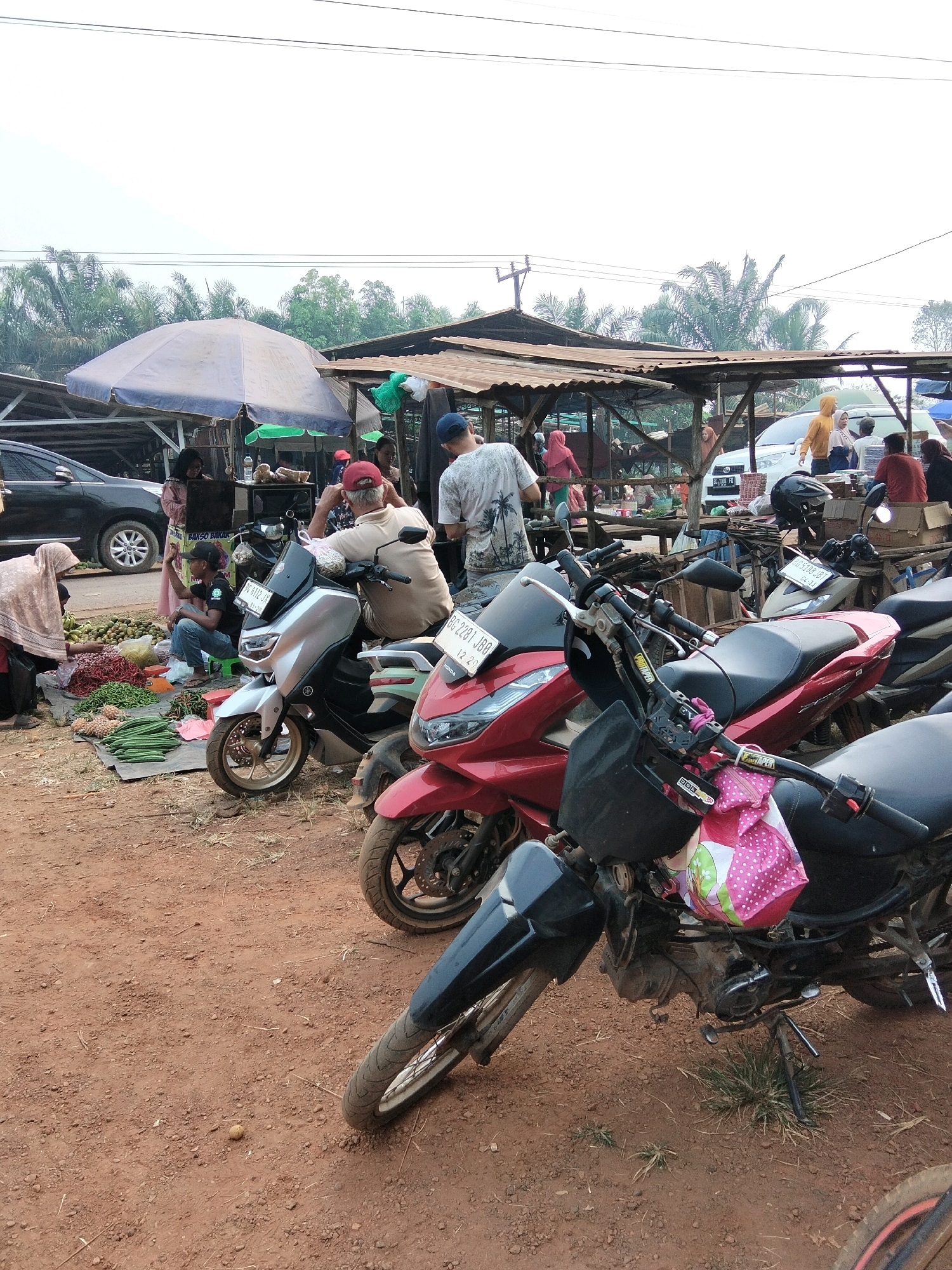 The image depicts an outdoor market scene in Tirto Sari, Indonesia. The foreground is dominated by several parked motorcycles, including a red scooter and a black and silver model. In the mid-ground, a variety of fresh produce, such as cucumbers and chili peppers, are laid out on mats on the ground. Several individuals are visible in the market, including vendors attending to their stalls and customers browsing the goods. Behind the market stalls, there are more people and vehicles, suggesting a bustling commercial area. The overall environment appears to be a typical Indonesian street market, with a mix of temporary structures and everyday life unfolding.