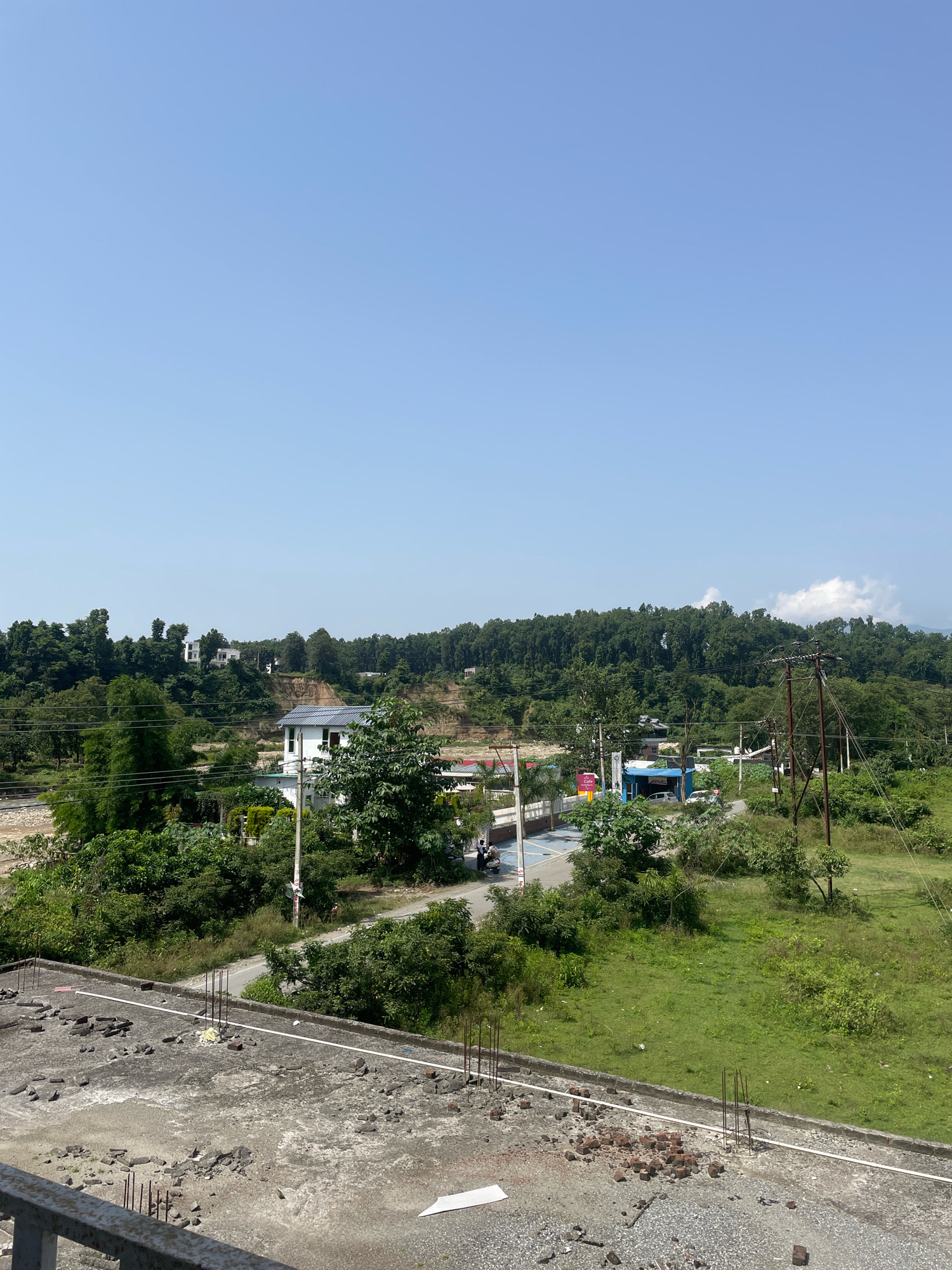 The image depicts a daytime scene in Kolhupani, India, with a clear blue sky dominating the upper portion of the frame. The foreground features a construction site with a concrete roof littered with rubble and exposed rebar, suggesting ongoing building work. A white pipe runs across the roof. Beyond this, a lush green landscape unfolds, characterized by trees, grass, and some residential buildings.
In the middle ground, a white building with a dark-tiled roof is visible, situated amidst dense greenery. A road runs past this building, with a few vehicles and pedestrians present. A small blue structure with a white roof is also visible near the road, possibly a shop or a café. Further in the background, rolling hills covered in dense forests rise towards the horizon. Several white houses are scattered on these hills, indicating a semi-rural or suburban setting. Power lines and utility poles are present, crossing the landscape and connecting various structures. The overall impression is one of a developing area with a blend of natural beauty and human construction.