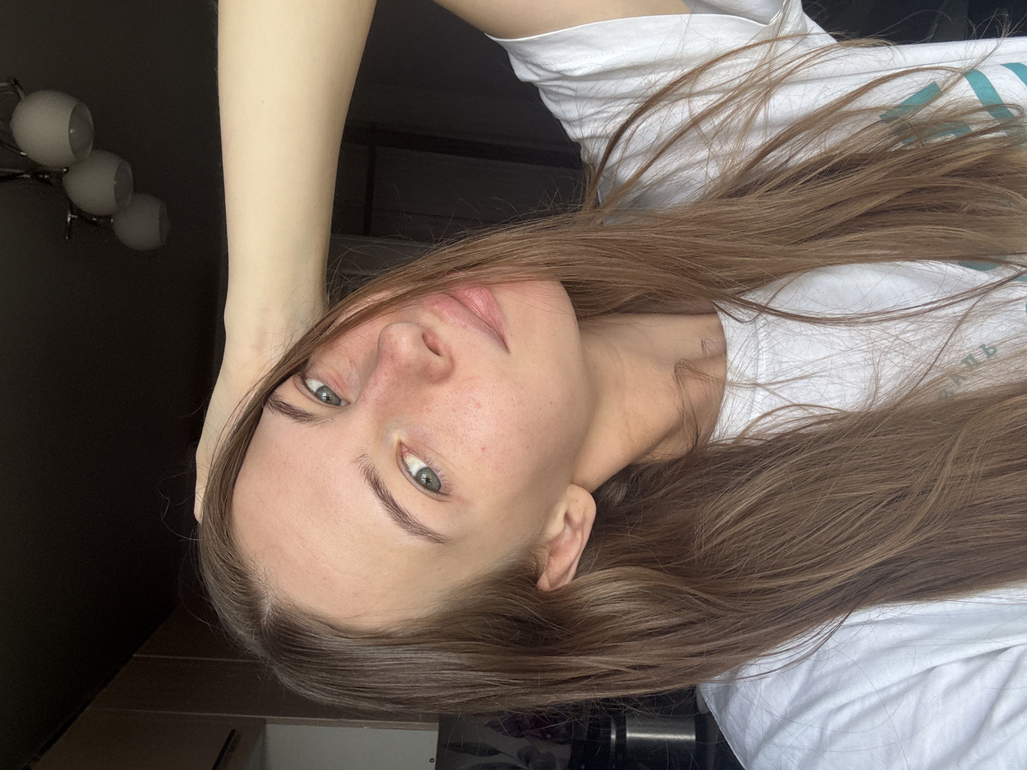 The image shows a close-up, waist-up, indoor shot of a young woman with long, light brown hair and light blue eyes. She is wearing a white t-shirt and has her right arm raised, with her hand resting on the back of her head. Her head is tilted to the left, and she is looking towards the camera. She has light skin with a few freckles on her nose and cheeks, and her lips are slightly parted. The background is dark and out of focus, with the hint of a light fixture on the ceiling. There are no visible signs of the city of Perm, Russia.