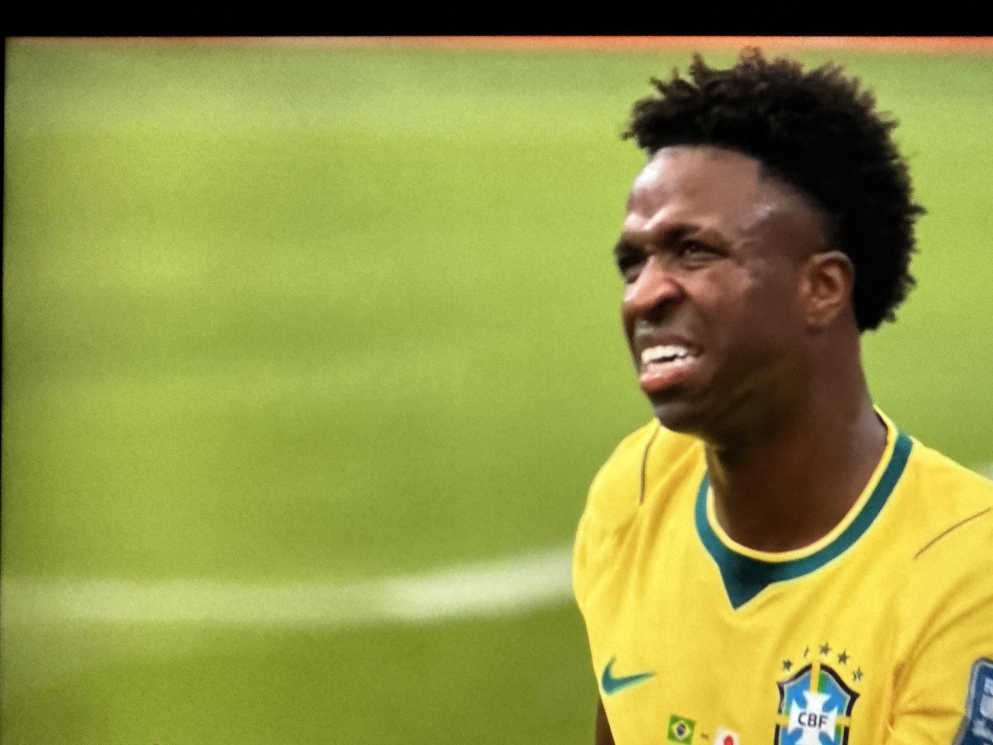This image shows a close-up of a Black male soccer player wearing a yellow Brazil national team jersey with green trim. He has dark, curly hair styled in a mohawk. The player's face is contorted in an expression of strain or exertion, with his mouth open and teeth visible. He is looking to his left. The background is a blurred green soccer field with a white line visible horizontally across the mid-ground. There is no indication of the location being Nha Trang, Vietnam.