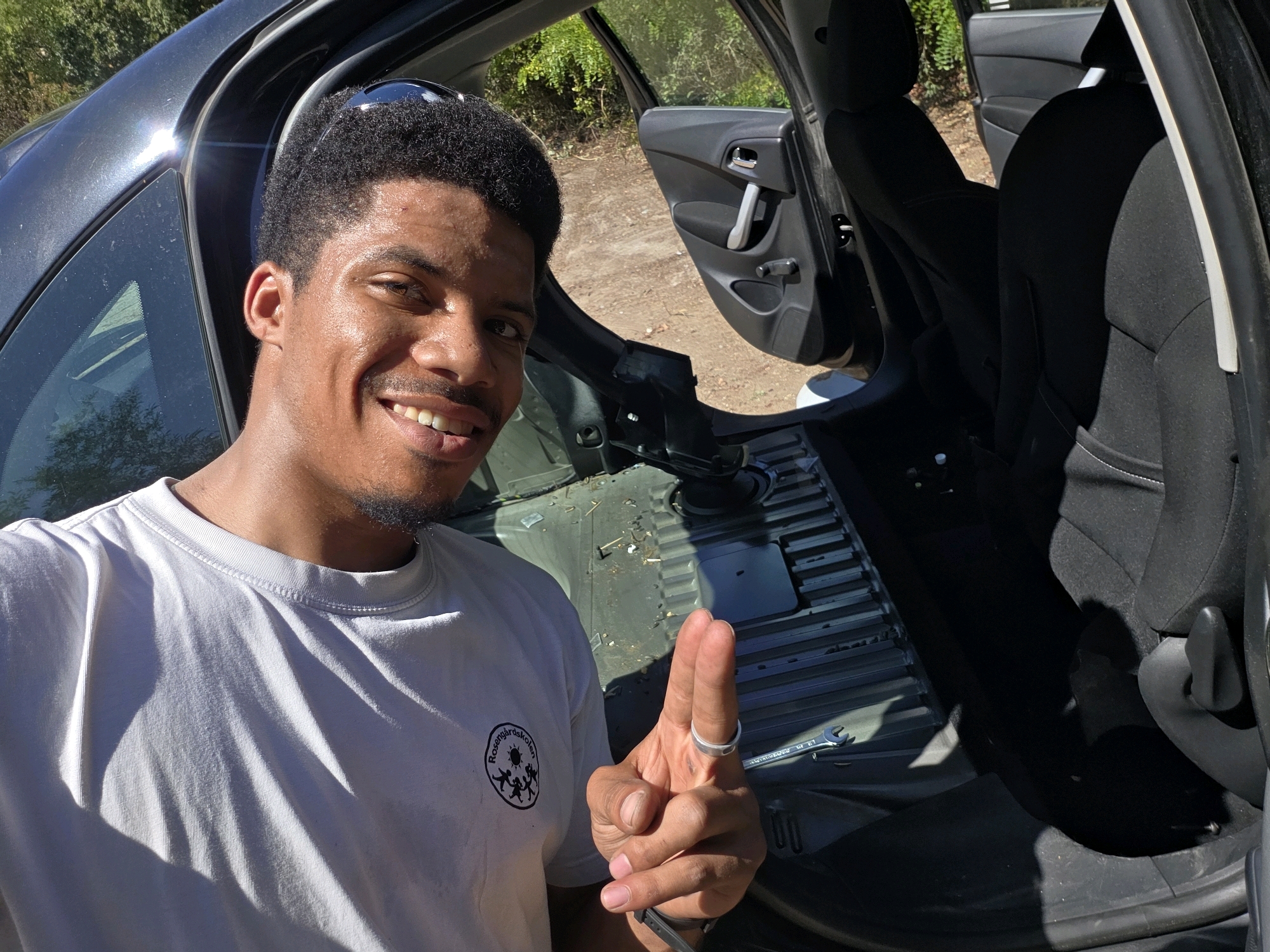A man with dark curly hair and a light complexion is standing next to the open door of a dark-colored car. He is smiling at the camera and wearing a light gray t-shirt with a circular logo on the left chest. His right hand is raised, with two fingers extended, and he is wearing a silver ring on his right index finger. The interior of the car appears to be partially disassembled, with some floor panels removed, revealing structural components and some debris on the floor. A wrench is visible on the floor to the right of the man. The background shows a dirt path with vegetation and trees, suggesting an outdoor, possibly rural or park-like setting. The lighting indicates it is daytime with bright sunlight casting strong shadows. The man is positioned in the foreground on the left side of the image, while the car interior and the outdoor environment are visible in the midground and background.