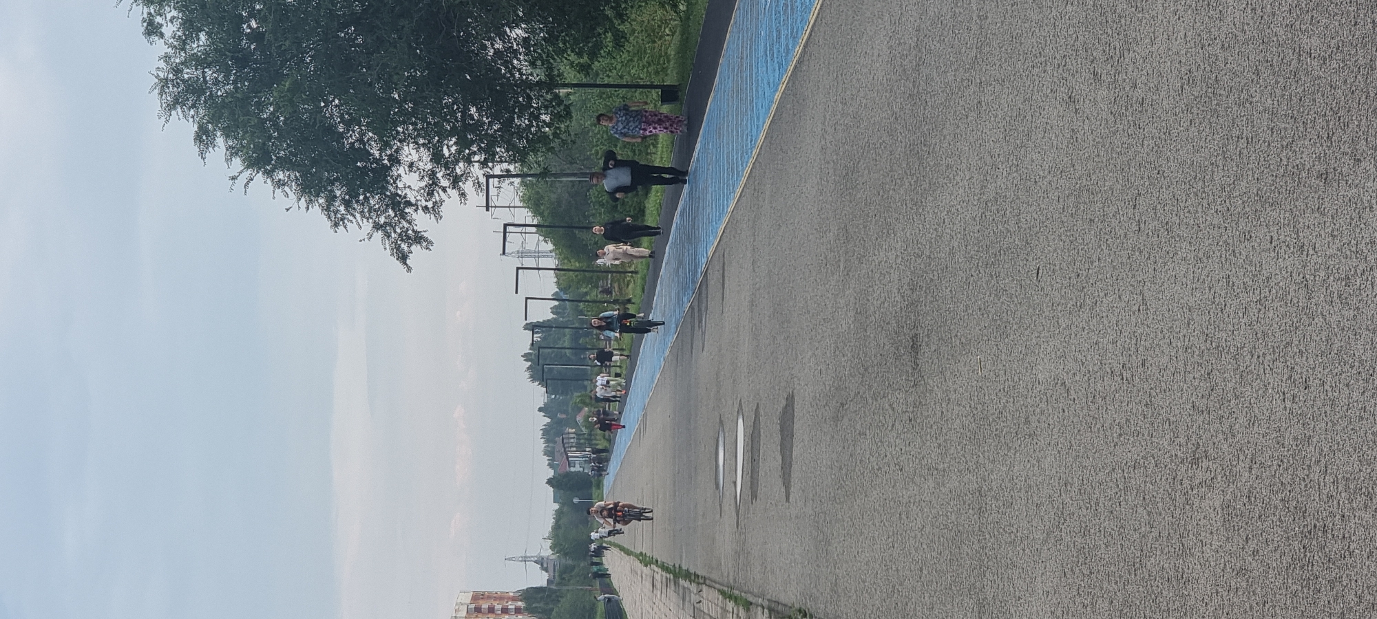 The image depicts a wide, paved pathway, likely a pedestrian and bicycle lane, in Aktobe, Kazakhstan. The pathway is flanked by a blue-lined section on the right, possibly a dedicated cycling track. A row of modern streetlights with dark poles and square heads runs along the right side of the blue section, spaced evenly apart. In the background, trees and greenery form a dense line, with a few buildings and utility poles visible beyond. The sky is overcast with pale clouds.
Several individuals are engaged in recreational activities on the pathway. On the left side, a person is riding a bicycle, and further up, another cyclist is visible. To the right of the blue-lined track, a group of people are walking. One man in a suit jacket and dark pants is walking towards the camera, while a woman in a patterned skirt and a dark top walks beside him. Further up the path, other individuals are walking and cycling. The pathway itself shows signs of recent rain with several puddles visible on the grey asphalt. The overall impression is of an urban park or recreational area designed for active use.