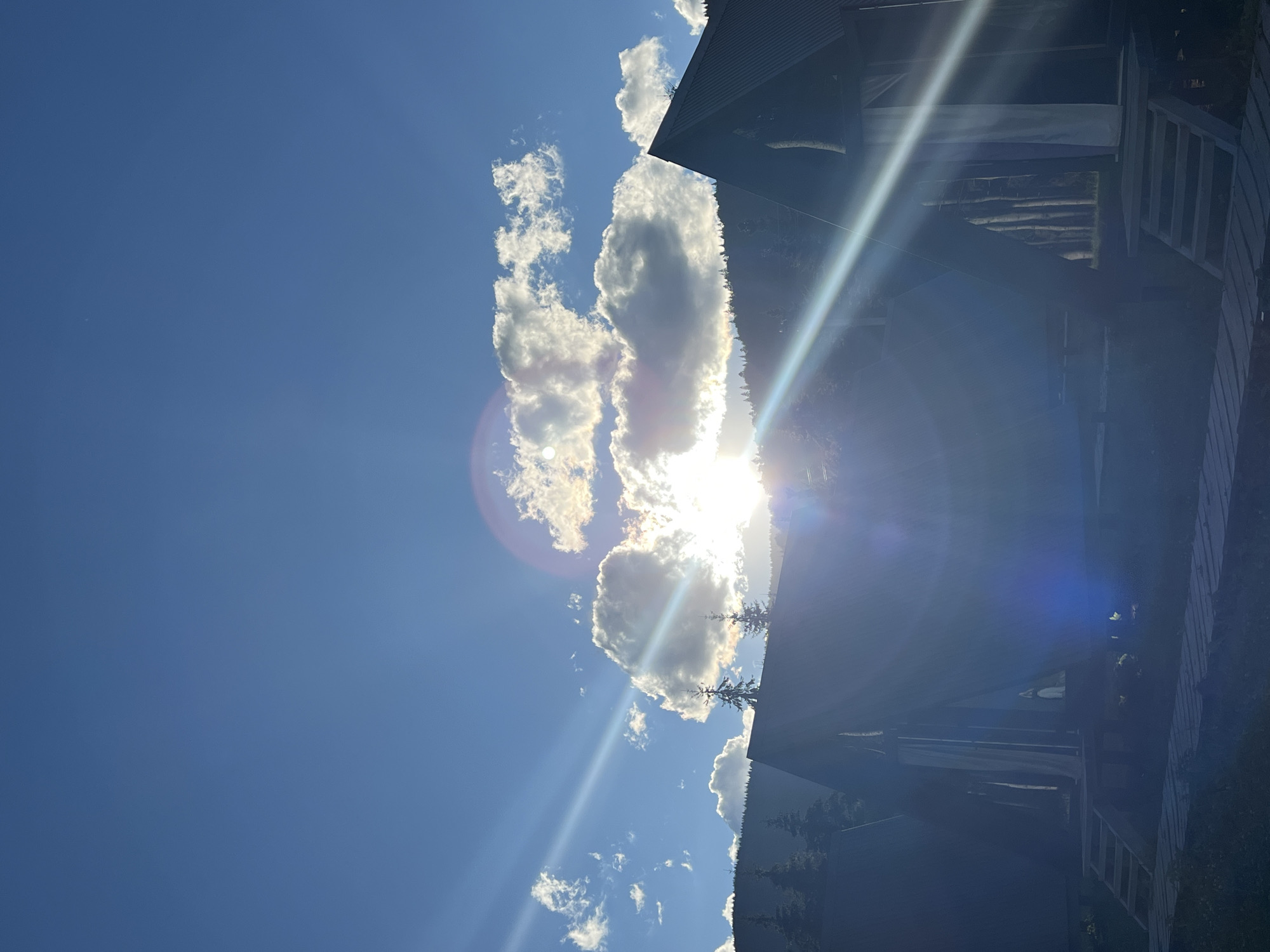 The image captures a sunny day in the city of Vorokhta, Ukraine, with a clear blue sky and scattered white clouds. The sun is shining brightly, creating lens flares and light beams that cut across the scene. In the foreground, there are several dark, triangular structures that appear to be glamping tents or small cabins, each with wooden stairs leading up to them. A wooden pathway is visible in front of the structures. Behind the cabins, a dense forest lines a hillside, with the tops of trees visible against the sky. The overall impression is one of a peaceful, natural setting.