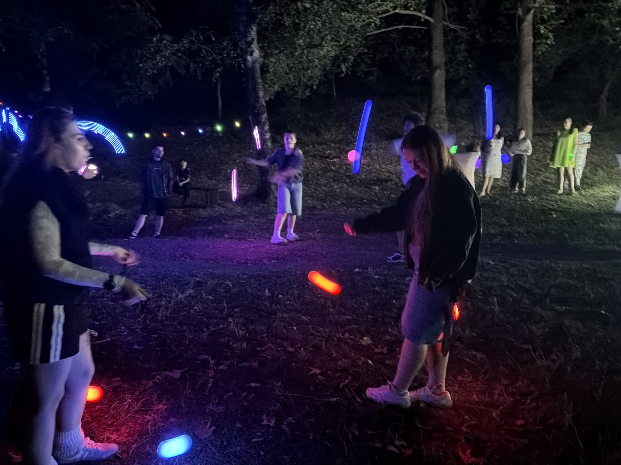 This image depicts an outdoor nighttime scene in the city of Zgorigrad, Bulgaria, where a group of people are participating in an activity involving illuminated objects. In the foreground, a person is visible from the waist down, wearing shorts with white stripes and white socks and sneakers. They are holding a glowing pink object in their right hand and appear to be in motion, possibly juggling or playing a game. Several other glowing objects, red and blue, are scattered on the ground around them, illuminating the dark, grassy terrain.
In the mid-ground, several individuals are engaged in similar activities. One person in the center is wearing a dark t-shirt and shorts and is actively juggling multiple pink glowing sticks. To their right, another person is holding a long, bright blue glowing stick. Further back, other individuals are standing and interacting, some holding illuminated objects, contributing to the vibrant, glowing atmosphere.
The background reveals a wooded area with trees, characteristic of the natural environment surrounding Zgorigrad. A string of colorful lights, likely decorative, is visible strung between trees in the upper left portion of the image, adding to the festive and illuminated nature of the event. The overall scene suggests a recreational or celebratory gathering taking place after dark.