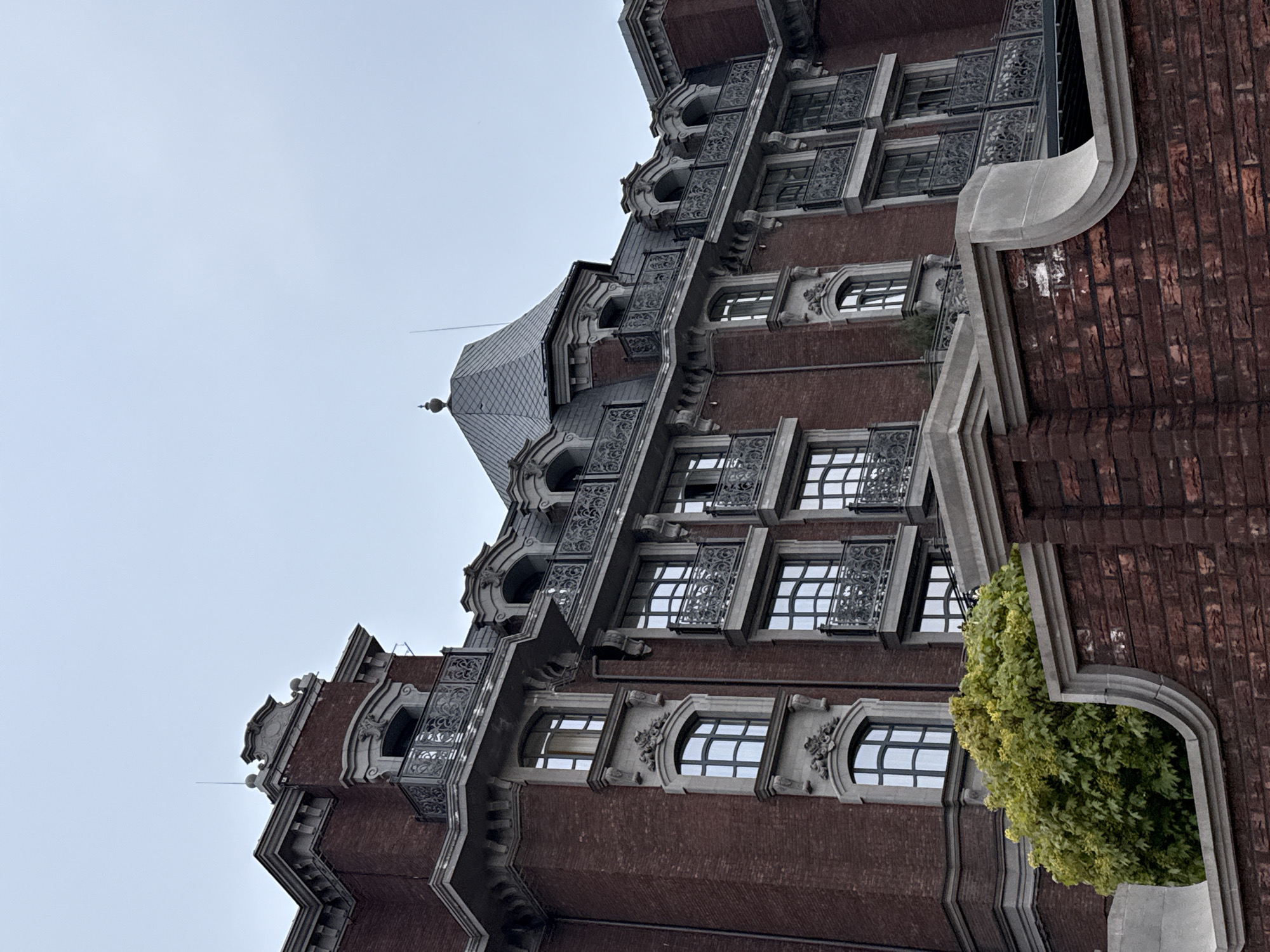 The image shows a partial view of a brick building with ornate stone or concrete detailing. The style is reminiscent of late 19th or early 20th-century architecture. There are multiple rows of windows, some with decorative ironwork balconies. A small, conical turret or tower is visible near the top. A tree or shrub is seen growing in an alcove on the side of the building. The sky is overcast. The building is located in Rostov-on-Don, Russia.