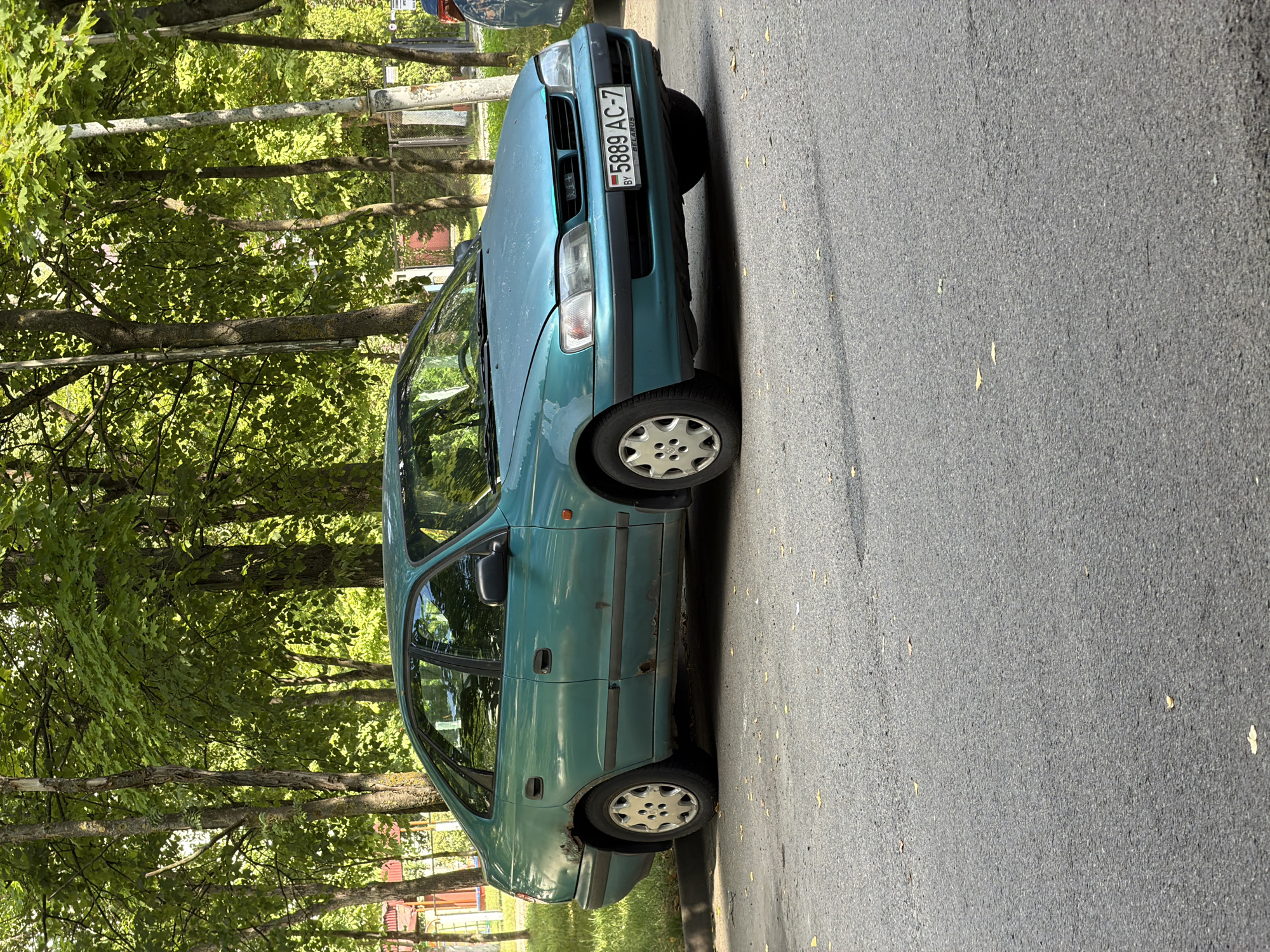 A teal-colored, four-door sedan is parked on a paved street in front of a row of trees. The car has a license plate that reads "5889 AC-7" and has "BY BELARUS" printed below it. The car shows signs of rust along the lower body panels and on the rear wheel arch. The street is asphalt and has some fallen leaves scattered on it. In the background, there are tall deciduous trees with green foliage. The lighting suggests it is daytime, possibly late morning or early afternoon.