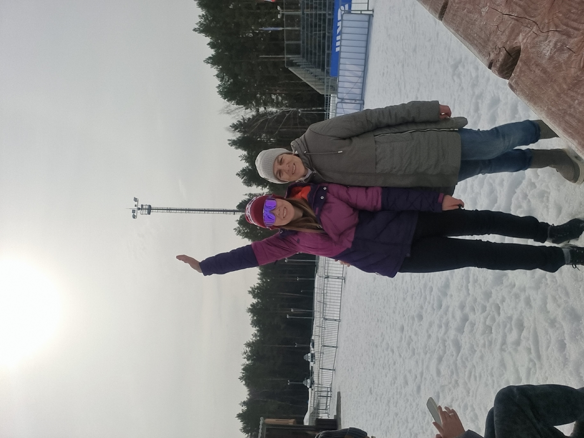 The image depicts an outdoor winter scene at a sports facility in Прикамский, Russia, under an overcast sky with a bright light source visible in the upper left. The ground is covered in snow.
Two adult females are positioned centrally. The female on the left wears a maroon knitted hat, purple-tinted mirrored ski goggles, a purple and fuchsia two-tone puffer jacket, black pants, and black boots. She has her right arm raised vertically, palm open, above her head. The female on the right wears a light gray knitted beanie, a gray puffer coat, blue jeans, and gray boots. She stands beside the first female, slightly behind her, with her right arm extended towards the other individual's waist. Both females are smiling.
In the background, a line of tall pine trees is visible. To the right, there are tiered spectator grandstands with blue lower sections. A tall metal floodlight tower stands in the center-right background. Metal barriers or fencing separate the foreground from a larger snow-covered area where several vehicles, possibly snowmobiles or utility vehicles, are parked further back to the left. A small wooden building is partially visible on the far left.
In the bottom left corner, a person's torso and hands are partially visible, holding a white smartphone. A ring is present on the left ring finger of this individual. In the bottom right corner, a section of a rough-hewn wooden structure, possibly a bench or table, is visible.