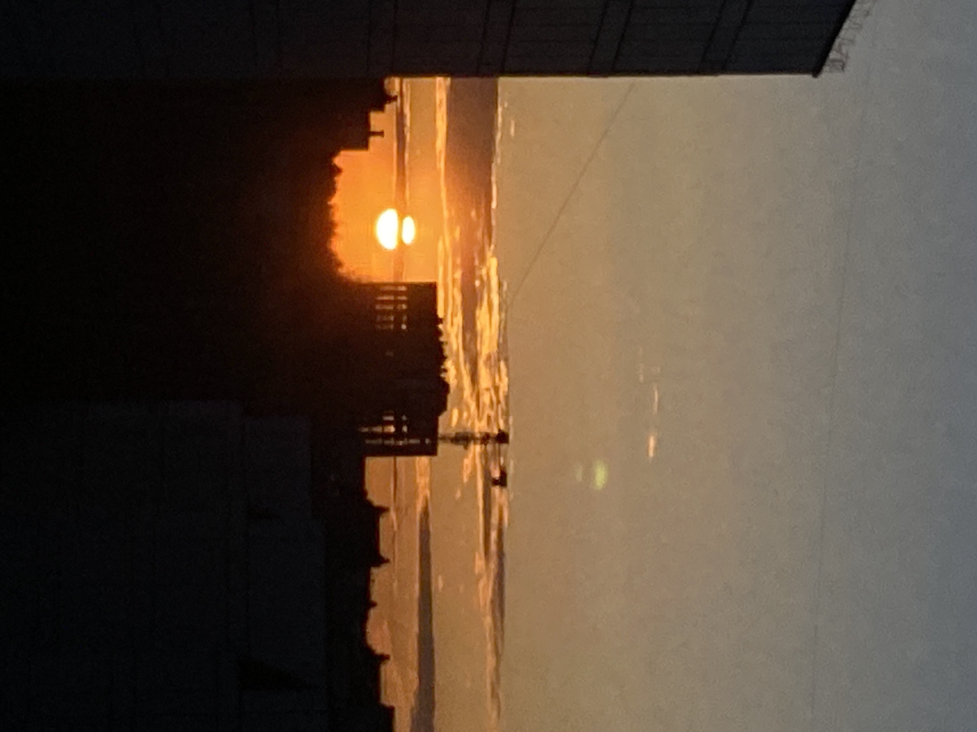 The image depicts a sunset over the Kama River in Chaikovsky, Russia. The sun appears as two distinct orbs due to atmospheric refraction, partially obscured by the silhouettes of buildings and trees on the riverbank. The sky transitions from a deep orange near the horizon to a lighter, greyish hue further up. The river reflects the sunset, creating a shimmering effect. A power line stretches across the upper right portion of the frame.