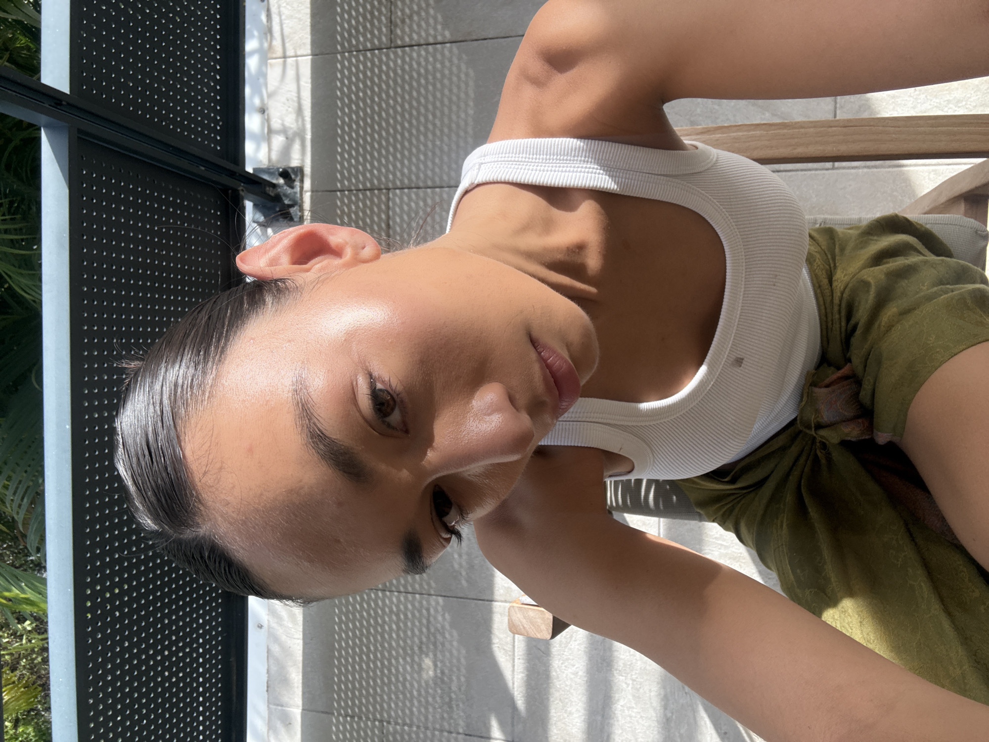 A person, likely a young woman, is captured in a sunlit, high-angle selfie taken outdoors. Her head is tilted, and she looks directly at the camera with a neutral expression. She has dark hair, slicked back, and prominent eyebrows and eyelashes. Her skin is highlighted by the bright, direct sunlight, particularly on her forehead, cheekbones, and shoulders.
She is wearing a white ribbed tank top and a flowing olive green garment, which appears to be a sarong or loose skirt. The background features a light-colored tiled floor or wall with strong, geometric shadows cast upon it, indicating intense daylight. To the left, a black perforated metal railing or fence is visible, alongside some green foliage. A wooden armrest, possibly part of a chair or bench, is also present. The scene suggests a warm, clear day in an outdoor setting, such as a balcony or patio in Saint-Martin, France. No text is visible in the image.