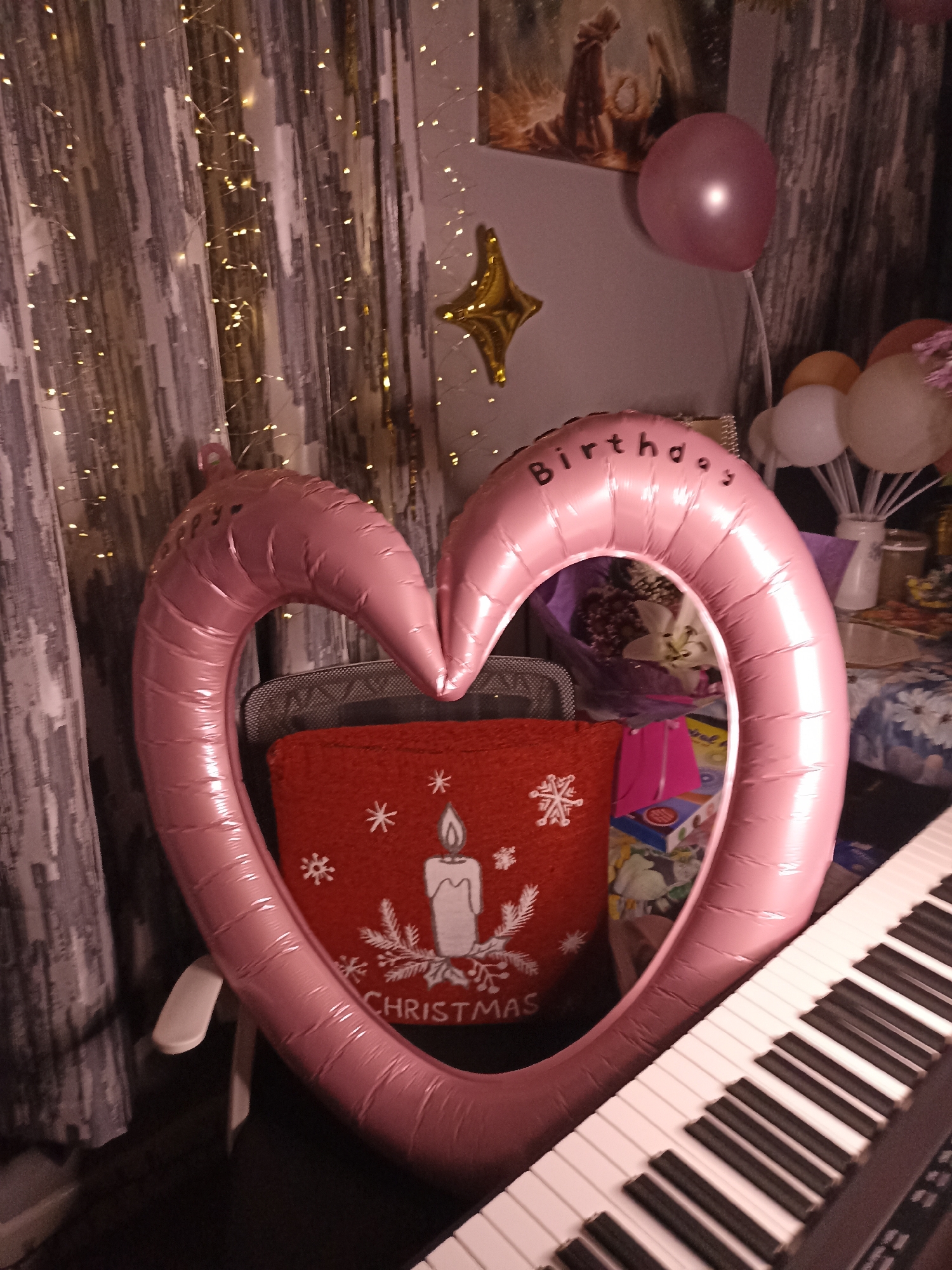 The image depicts an indoor scene, likely a room decorated for a celebration. A large, pink, heart-shaped balloon with the word "Birthday" printed on it is prominently displayed. To the right, a portion of a keyboard is visible. Behind the heart balloon, a red cushion with a Christmas-themed design, featuring a candle and snowflakes, is placed. In the background, there are curtains adorned with fairy lights and other balloons, including a pink one and some in white and orange. A framed picture hangs on the wall above the pink balloon. There is no indication of the location being Annagry, Ireland, or any specific city. The image does not contain any people.